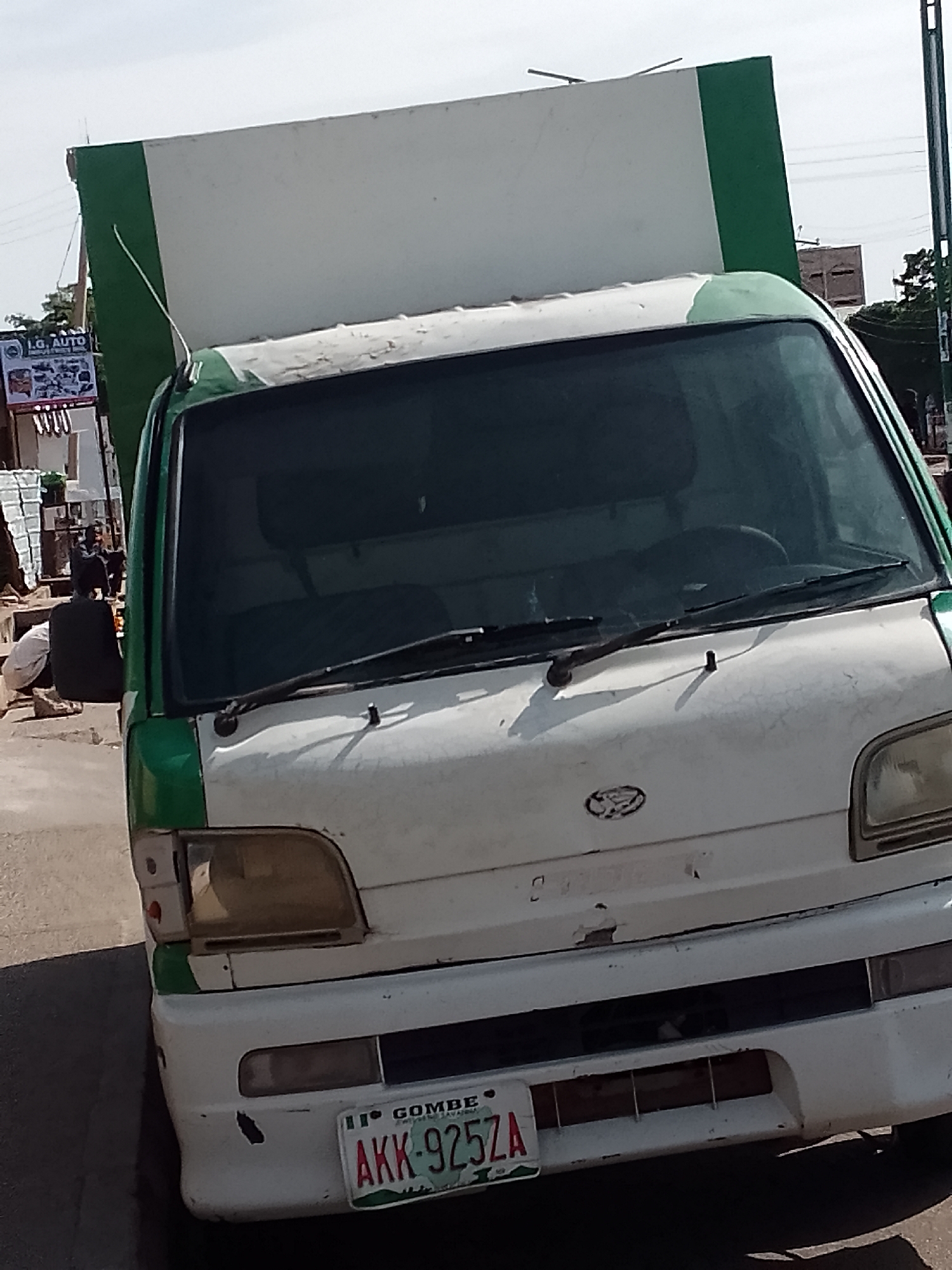 An up-close, eye-level, medium shot captures a white and green delivery truck parked on a paved street, likely in Pantame, Nigeria. The truck occupies the majority of the frame, with its front grille, headlights, windshield, and the side of its cargo bay visible. The vehicle appears weathered, with some rust and peeling paint. A license plate on the lower front of the truck reads "GOMBE AKK-925ZA". In the background, partially obscured by the truck, is a glimpse of a billboard advertising "I.G. AUTO", and a person, dressed in a black top and light-colored trousers, is crouched down on the left side of the image. The sky above is bright and clear, suggesting daytime. The overall impression is of a busy street scene in a Nigerian city.