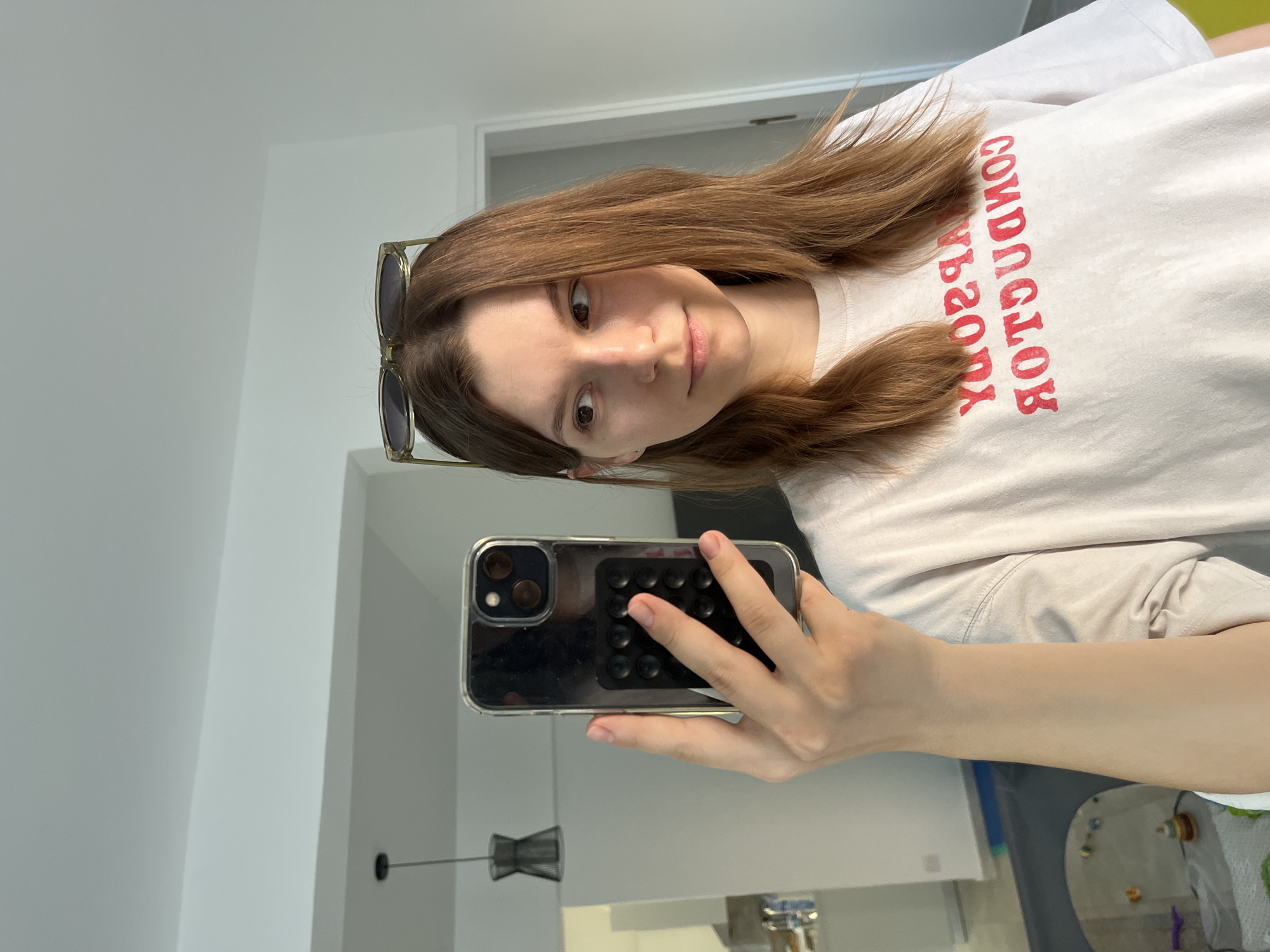 In an indoor setting, a woman with light brown hair stands in front of a mirror, taking a selfie with her phone. She is wearing a white t-shirt with red lettering that reads "CONDUCTOR" in reverse. Sunglasses are perched on top of her head. The background reveals a hallway with a light-colored door and a glimpse of a living area with furniture. The room is brightly lit, suggesting it is daytime. The overall scene depicts a casual, everyday moment.