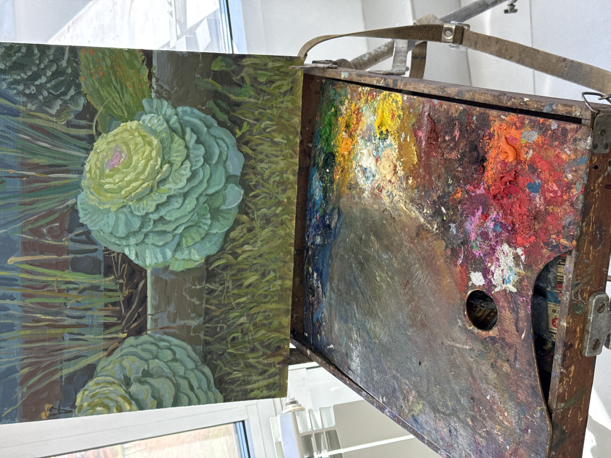 The image shows a painter's setup with a painting on the left and a palette on the right, both presumably resting on an easel. The painting depicts green plants or cabbages in an impressionistic style. The palette is heavily used and covered in a multitude of colors, including yellows, reds, blues, and grays, with a hole in the center. A canvas strap and easel mechanisms are visible above. A white framed window sits behind the easel. The location of Izhevsk, Russia cannot be confirmed from the visual information provided.