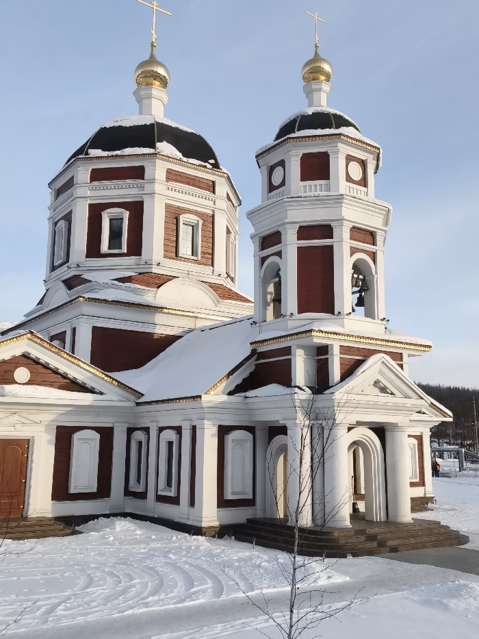 The image displays a Russian Orthodox church located in the city of Im. Gastello, Russia, under clear winter conditions. The church structure, painted in reddish-brown with prominent white architectural trim and gold accents, features a main building with an octagonal drum topped by a black dome, a golden onion cupola, and a golden Orthodox cross. To its right, a two-tiered octagonal bell tower rises, similarly crowned with a black dome, a golden cupola, and a cross. The bell tower's upper tier contains open arched windows revealing internal bells.
Snow covers all horizontal roof surfaces of the church. The ground surrounding the edifice is entirely blanketed in white snow, with tire tracks or similar patterns visible in the immediate foreground. A small, bare deciduous tree stands in the snow in the lower foreground. The sky is clear and bright blue. On the far right, in the background, a person wearing an orange jacket is partially visible, standing on a snow-covered path near a distant line of evergreen trees. Another indistinct figure is further back. Two entrances are visible: one dark brown door on the left side of the main structure and a lighter, recessed entrance with an arched portico and white columns on the right side of the building's base.