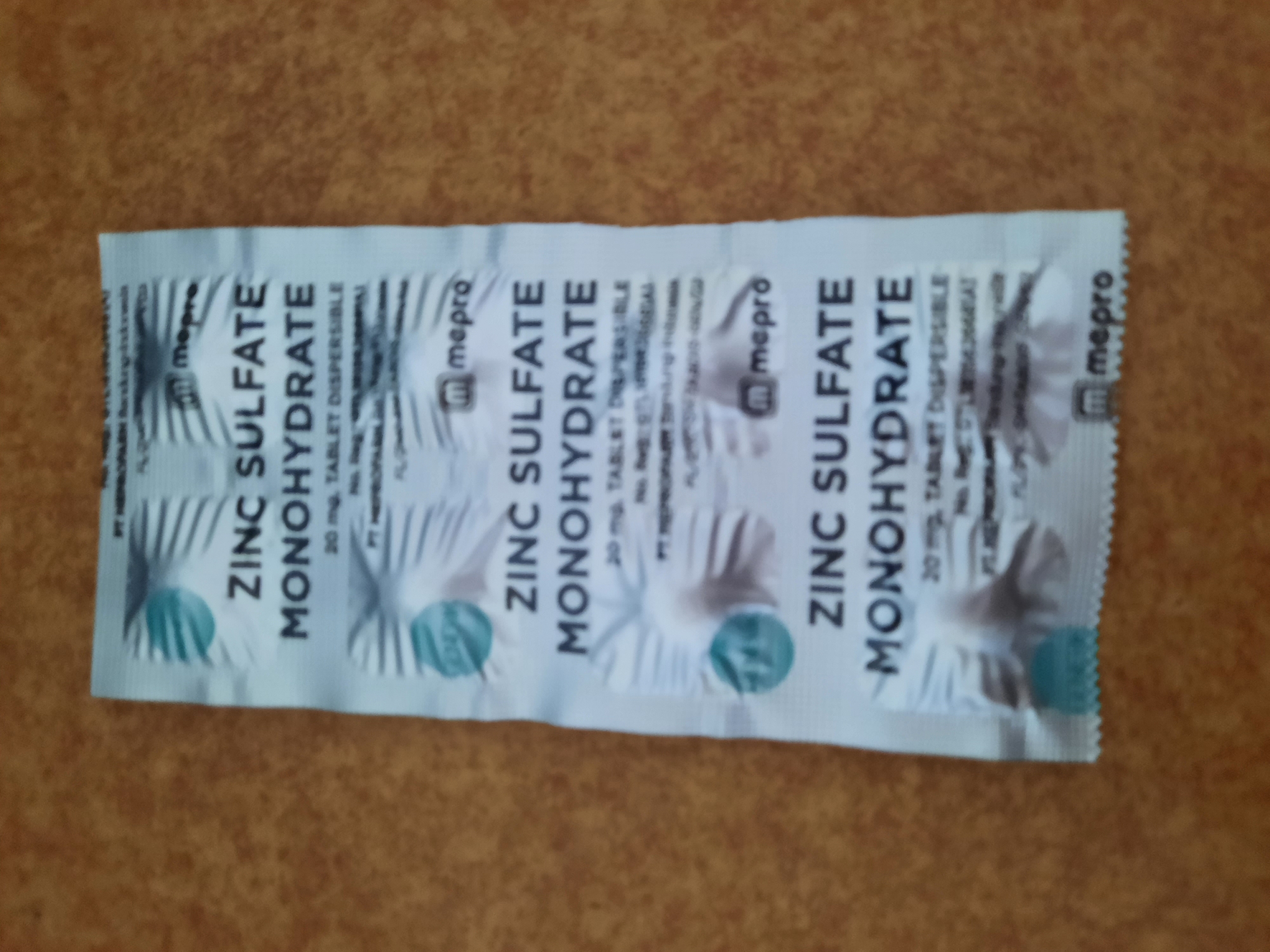 The image shows a blister pack of "ZINC SULFATE MONOHYDRATE 20 mg, TABLET DISPERSIBLE" medication. The blister pack is white with blue and gray graphics. It appears to contain three individually sealed compartments, each with the same branding and medication information. The overall condition of the packaging suggests it is a consumer product.
The background is a textured surface, likely a wooden table or countertop, with a warm brown tone. The lighting is somewhat dim, casting soft shadows. The image is taken from a slightly overhead angle.
While the image shows a product that could be purchased in Lhokseumawe, Indonesia, there is no visual information within the image itself that directly indicates its location in that specific city. The product's branding, "PT MEPROFARM," is visible, which is an Indonesian pharmaceutical company, but this does not pinpoint the location to Lhokseumawe specifically.