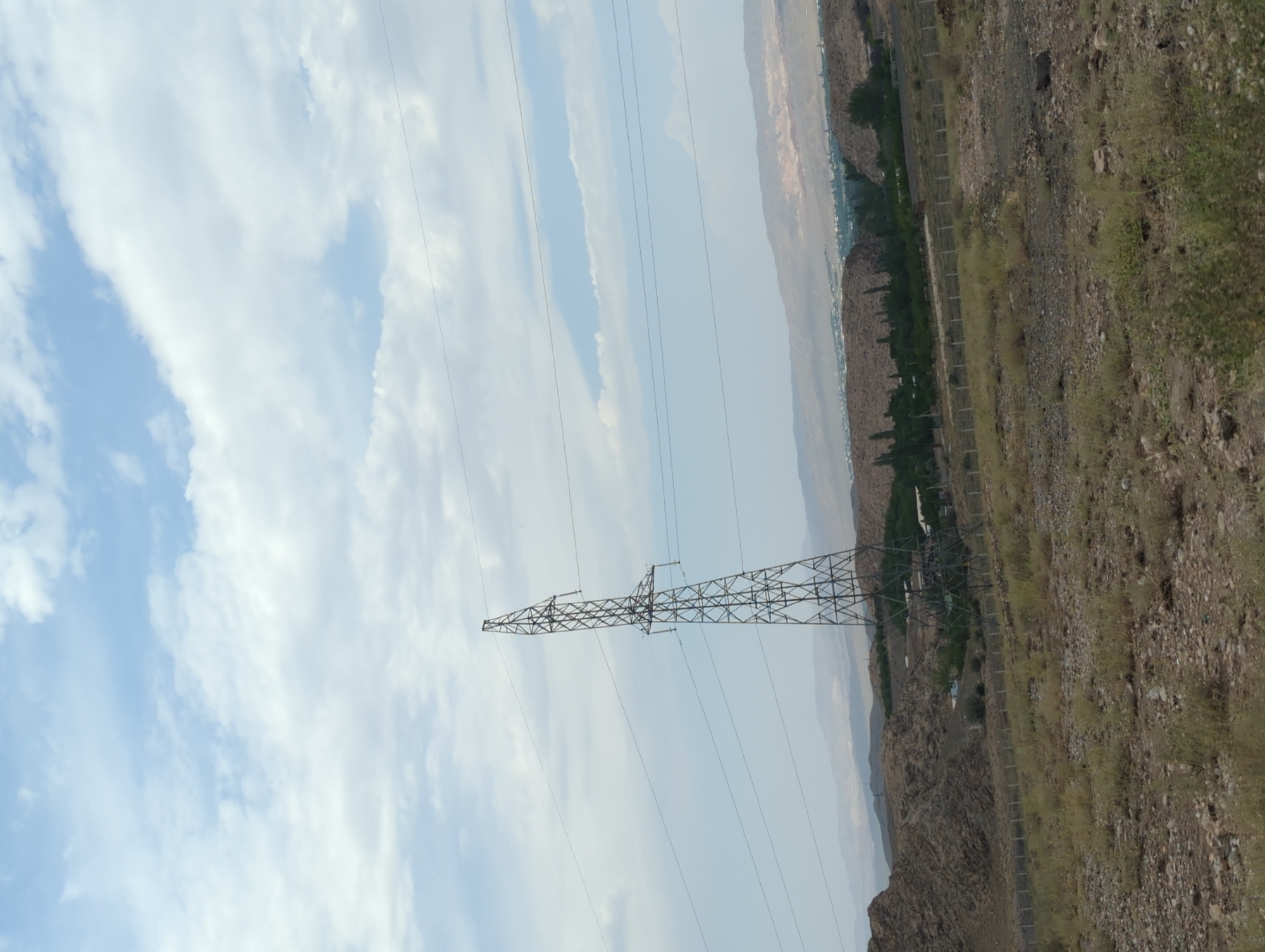 A wide shot captures a vast, arid landscape under a partly cloudy sky. Dominating the mid-ground is a tall, metallic power line pylon, its lattice structure stark against the pale blue and white of the clouds. Several power lines extend horizontally from the pylon, disappearing into the distance. The terrain is rugged and dry, with rocky hills and sparse vegetation in the foreground and mid-ground. In the distance, a more settled area with trees and buildings is visible, suggesting a small settlement or village. The background features layered, hazy mountains. The scene appears to be located in a rural or semi-arid region.