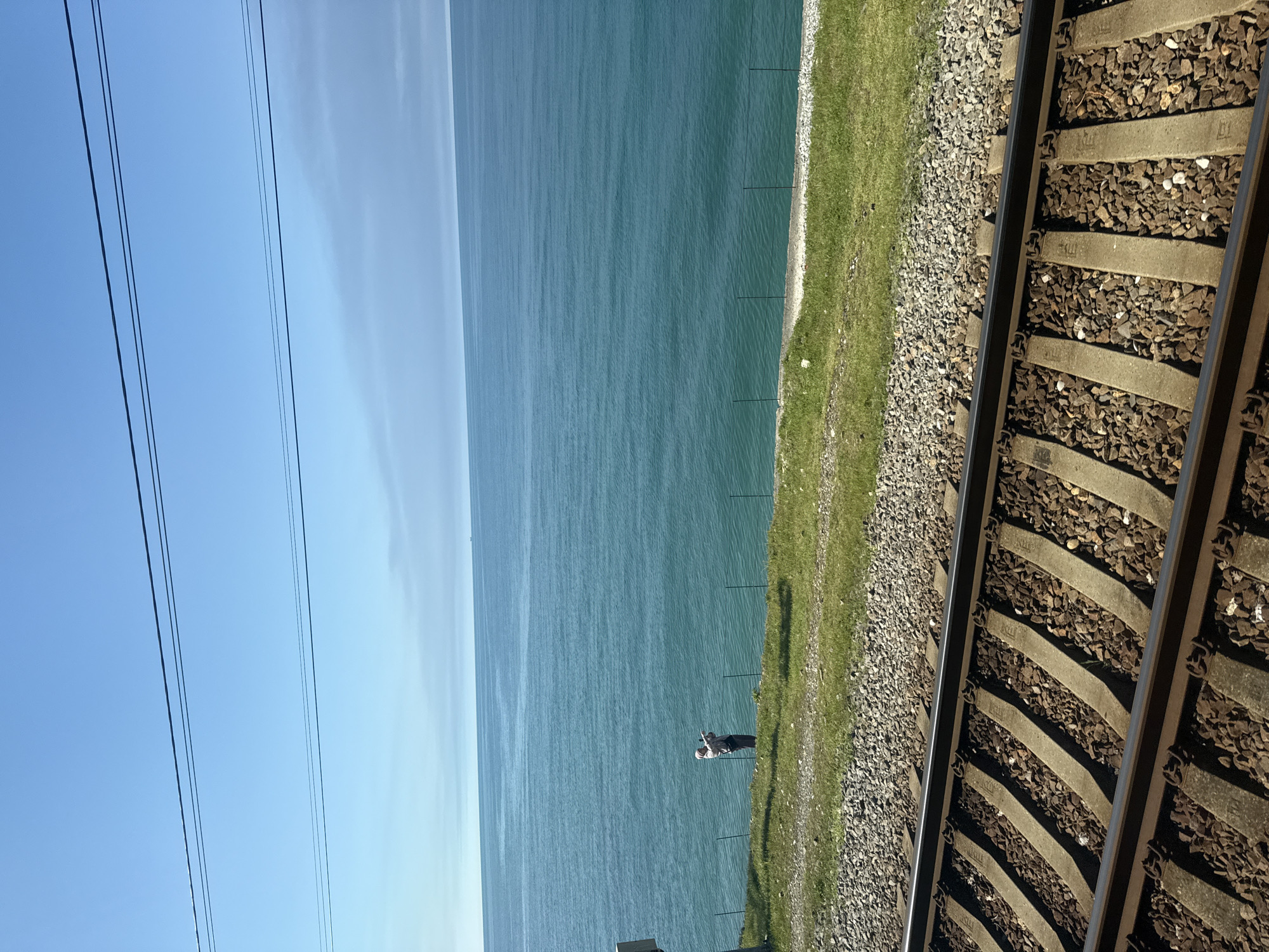 Here is a detailed description of the image:
The image presents a landscape dominated by the sea on the left and a railway track on the right. The sky is a clear blue.
A strip of green grass runs parallel to the sea and the railway track, separating them. A person is standing between the grass and the sea and appears to be taking a photo. Fences are seen between the grass and the sea.
Power lines run along the left side of the image near the blue sky.
The railway track occupies the right side of the frame and extends from the foreground into the background. It is composed of rails, sleepers, and ballast.