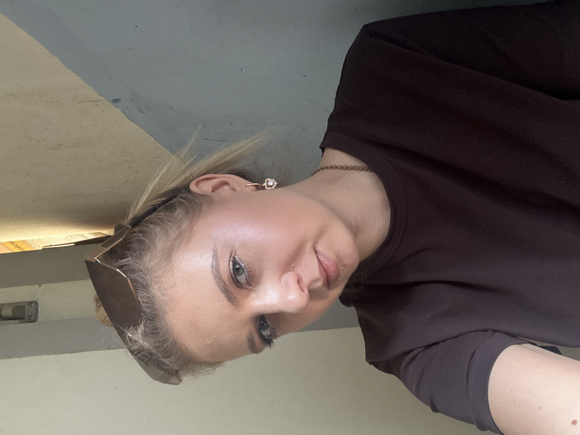 A young woman with blonde hair pulled back and sunglasses perched on her head takes a selfie. She is wearing a dark brown t-shirt and a delicate gold necklace with a charm. She has a small stud earring in her visible ear. The background appears to be a textured wall with a distinct diagonal color change from light yellow to gray-blue. A light fixture is visible on the wall to the left. The lighting suggests an indoor setting, possibly a hallway or corner of a room.