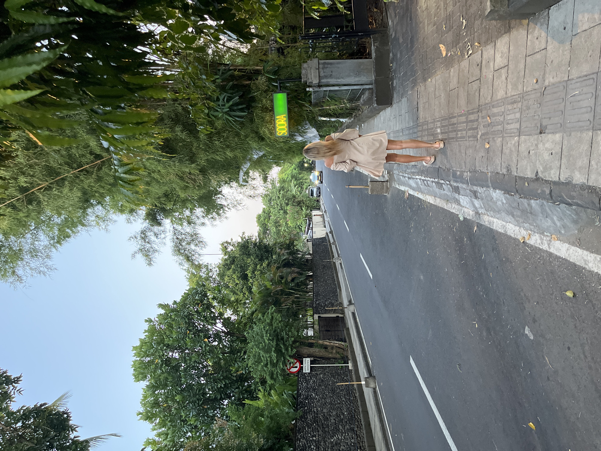 The photograph, taken in Unknown, Indonesia, presents a street scene with a woman as the primary subject. The woman, with blonde hair, is dressed in a light-colored dress and sandals. She is walking on a sidewalk. A road runs parallel to the sidewalk. The road has white lines marking the lanes and shows some vehicular traffic in the distance. Lush green trees line both sides of the street, creating a canopy overhead. There is a sign indicating the name "SCOORA," appearing to be illuminated in green. A no-entry traffic sign is visible, partially obscured by foliage, along the side of the road. The sidewalk is constructed of stone or concrete pavers, featuring a textured strip. The lighting indicates either early morning or late afternoon.