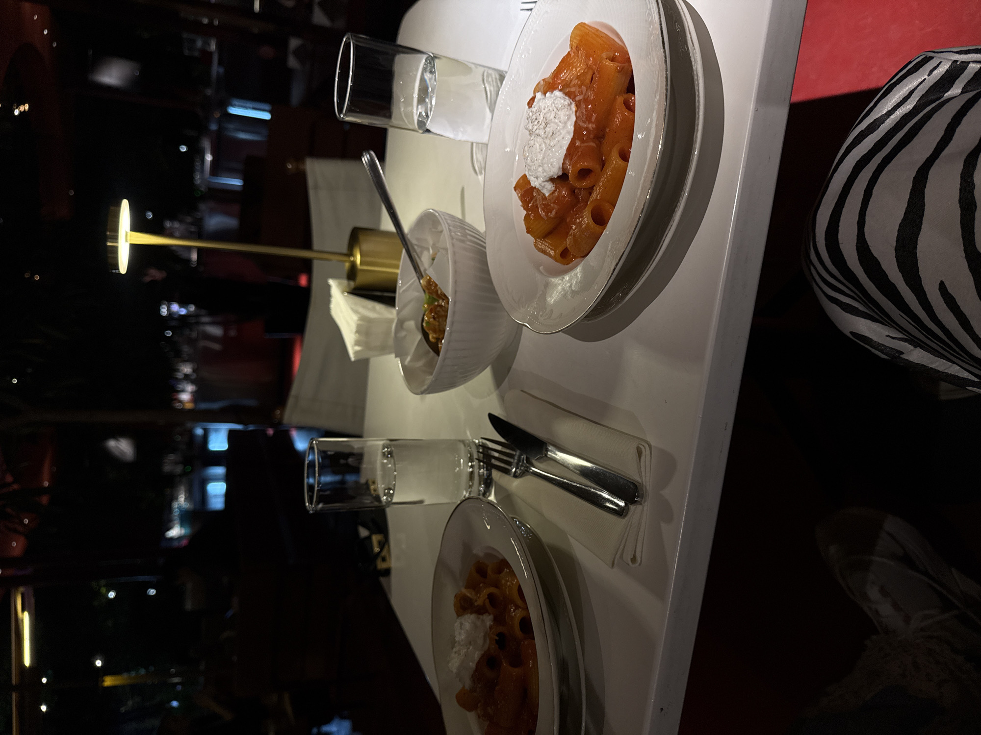 A high-angle shot depicts a table setting at a restaurant, likely in Yerevan, Armenia, given the context clues. The tabletop is white and oriented vertically in the frame. Two plates of pasta are visible, each featuring rigatoni in red sauce, topped with a dollop of white cheese. One plate is near the top center, while the other is at the bottom. A small white bowl, containing a brown food item, sits between the two plates. Silverware, including forks, knives, and spoons, is neatly arranged near the plates. Two glasses of water are present. One glass is next to the top plate, and the other is next to the bottom plate. In the background, a lamp with a gold stand is partially visible. To the right of the top plate is a zebra-patterned object, possibly a piece of clothing or a seat. The background is dark, suggesting an indoor setting with dim lighting, possibly at night. The restaurant appears upscale, with a minimalist aesthetic.