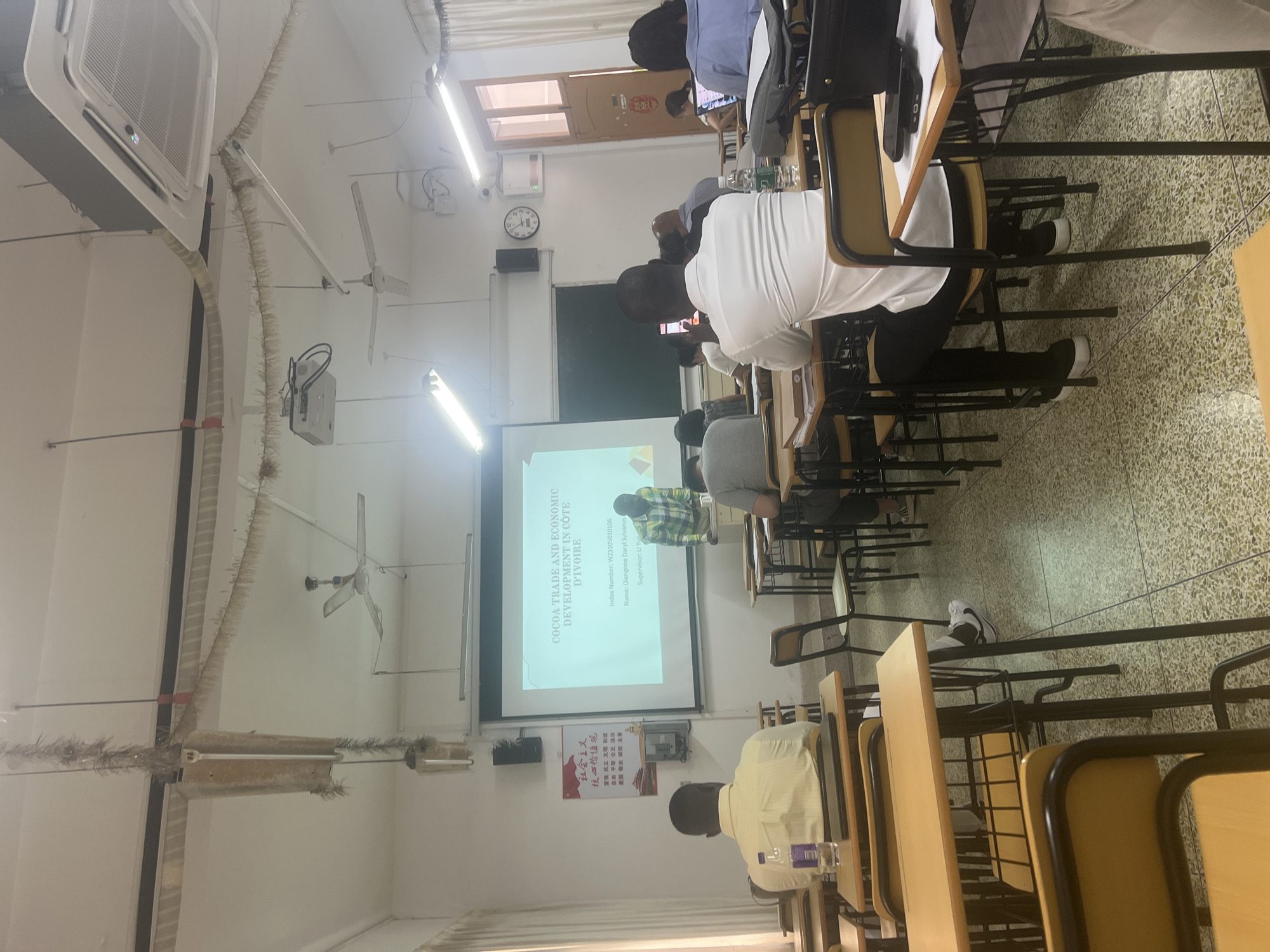 The image shows a lecture hall with students seated at desks facing a projector screen. The screen displays text related to "COCOA TRADE AND ECONOMIC DEVELOPMENT IN COTE D'IVOIRE". A presenter, a Black male wearing a checkered shirt, stands near the screen. Several students, predominantly Black males, are visible in the foreground and middle ground. One student in the foreground is using a mobile phone. The room features desks, chairs, fluorescent lighting, ceiling fans, and an air conditioning unit. There are also Chinese characters on a poster on the wall. The floor is tiled with a speckled pattern. The overall scene depicts an academic setting, likely a university lecture or presentation.