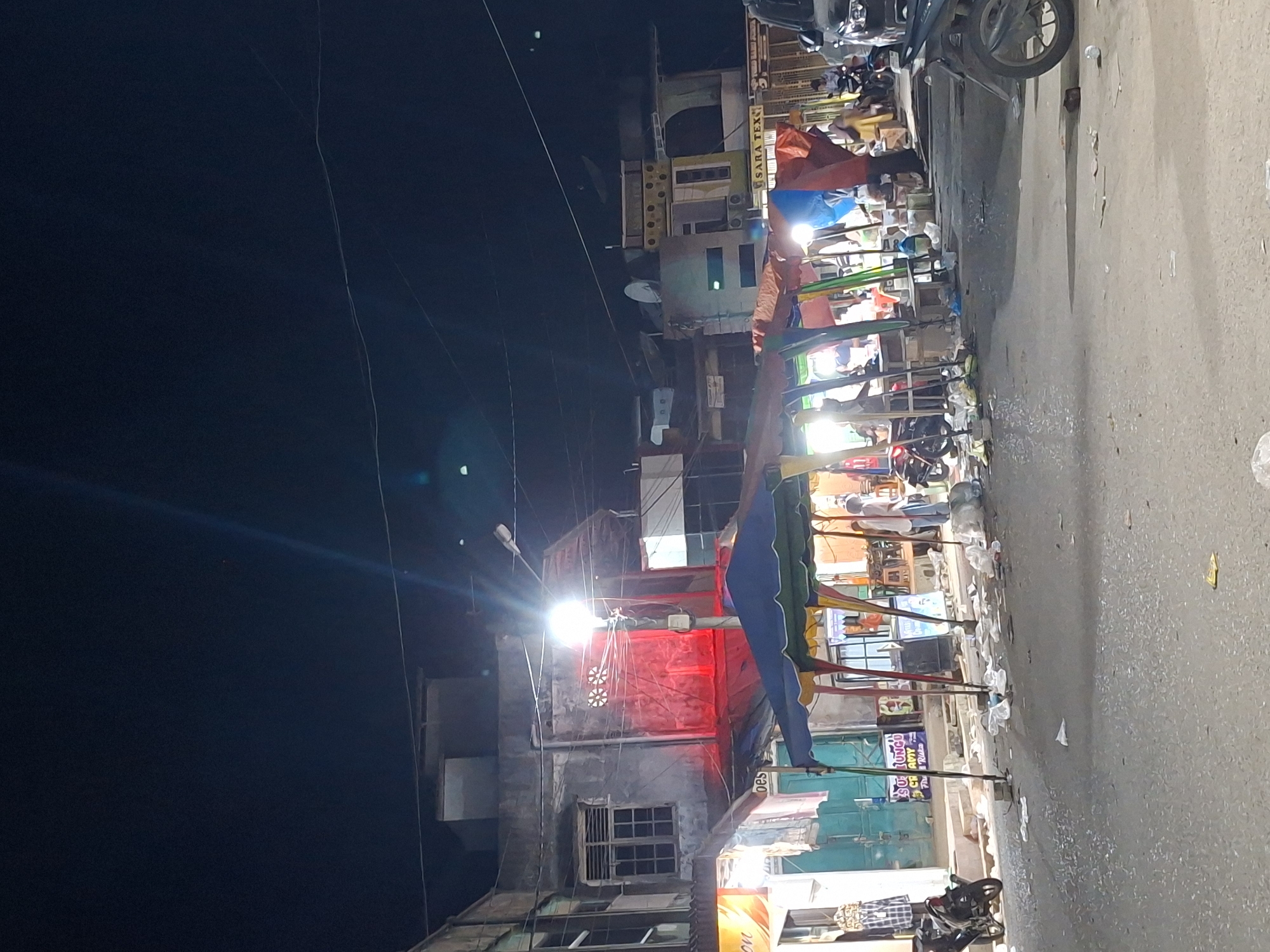 The scene depicts a street in Keudegeudong, Indonesia, at night. Street vendors are set up along the roadside. The street is littered with trash. A building with a red facade is visible in the background. The sky is dark, and power lines are visible. A motorcycle is parked nearby. People are present near the stalls and under the light fixtures. Banners are placed along the stalls.