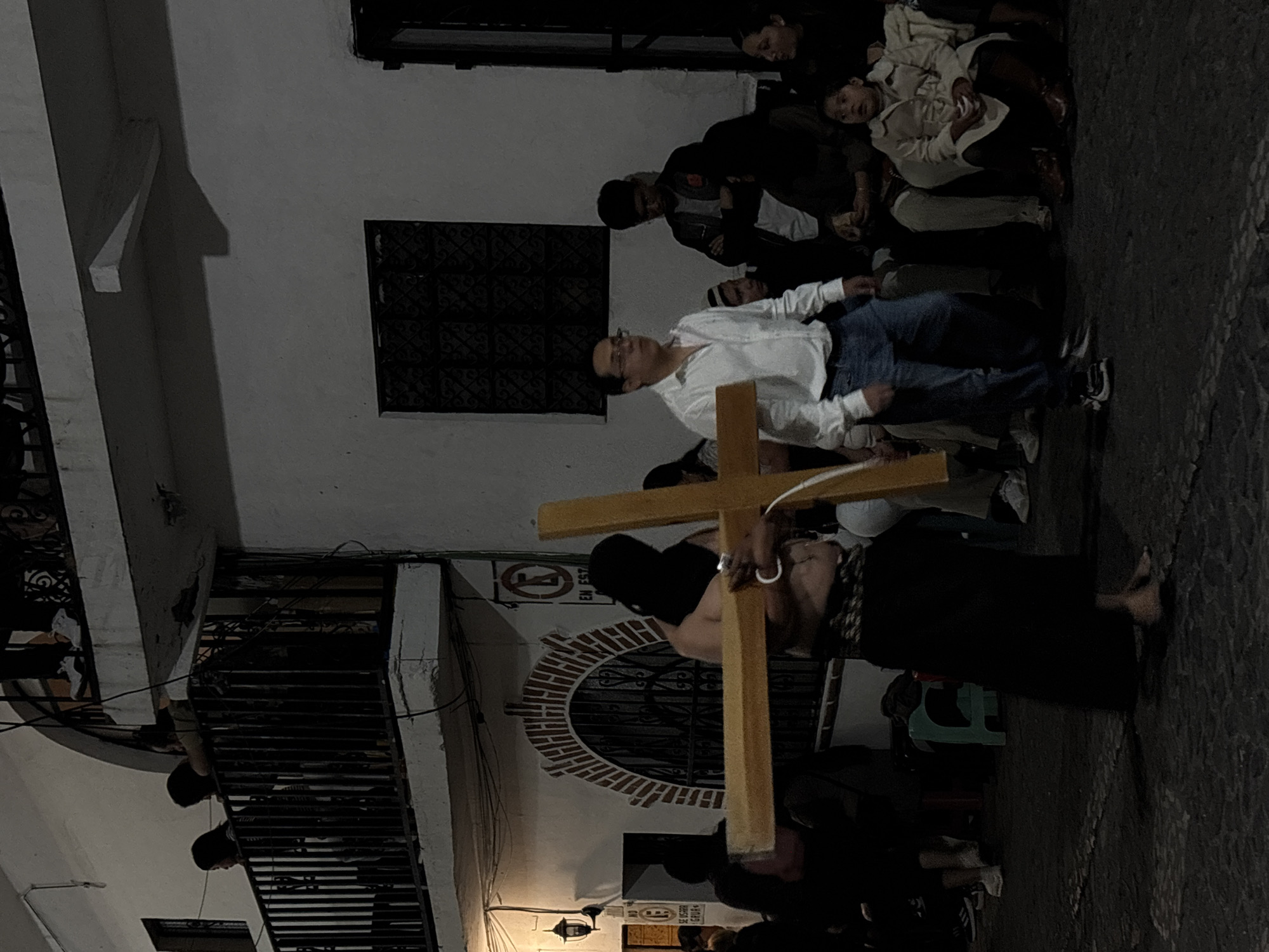 The image captures a religious procession at night in Taxco, Mexico. A barefoot man carries a large wooden cross through a cobblestone street. He is wearing a dark robe and a hood or head covering. A crowd of people watches, some dressed casually. Buildings with white walls and iron balconies line the street. A window with a decorative wrought iron grill is visible in the background. Streetlights illuminate the scene.