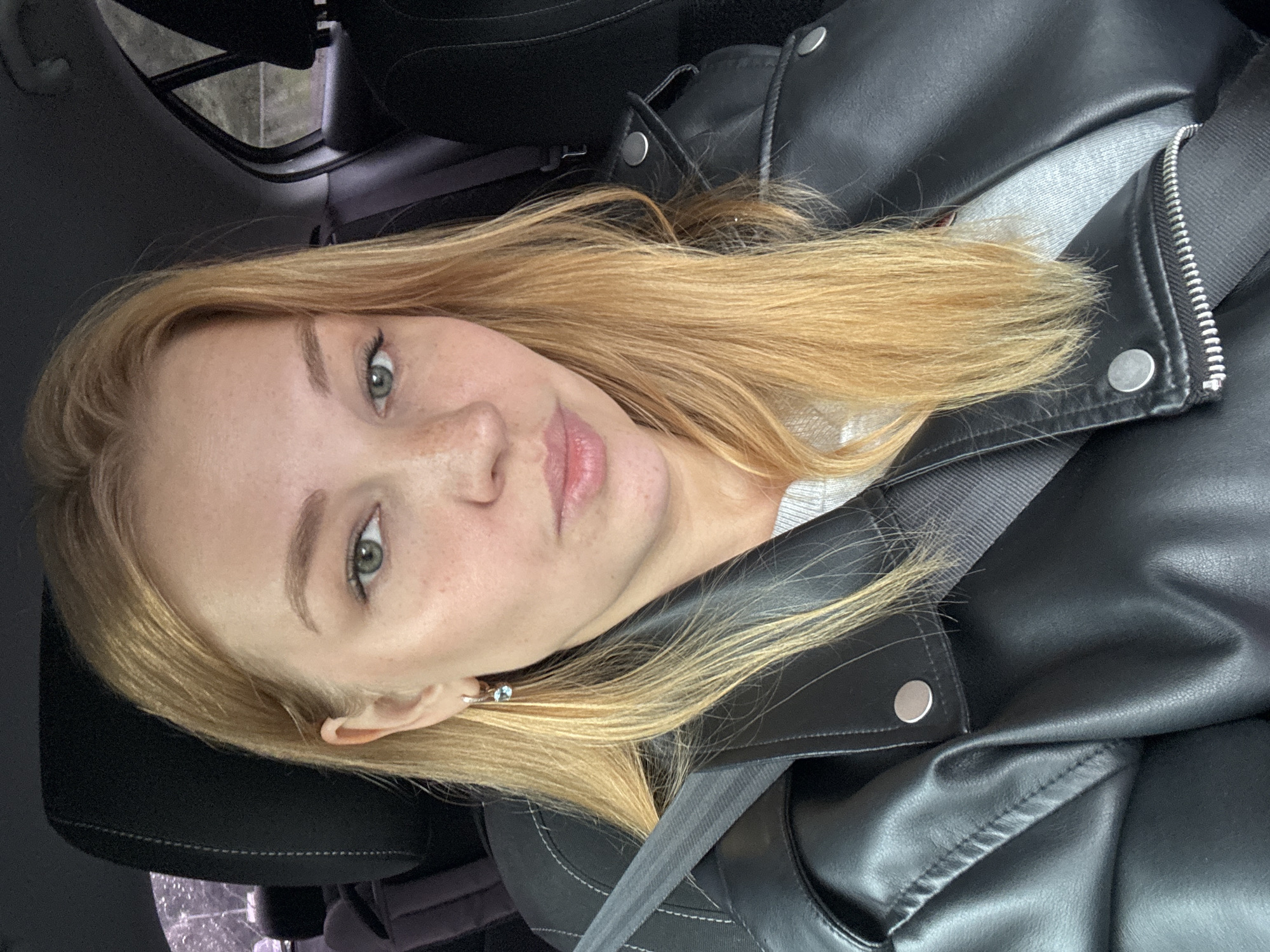 A young Caucasian woman with long blonde hair and fair skin with freckles is in the driver's seat of a car. She is wearing a black leather jacket over a grey sweatshirt and a seatbelt. The background shows the interior of the car, including the headrest, door, and a rainy outdoor scene through the window. The image appears to be a selfie taken by the woman. The location is identified as Rusanovo, Russia.