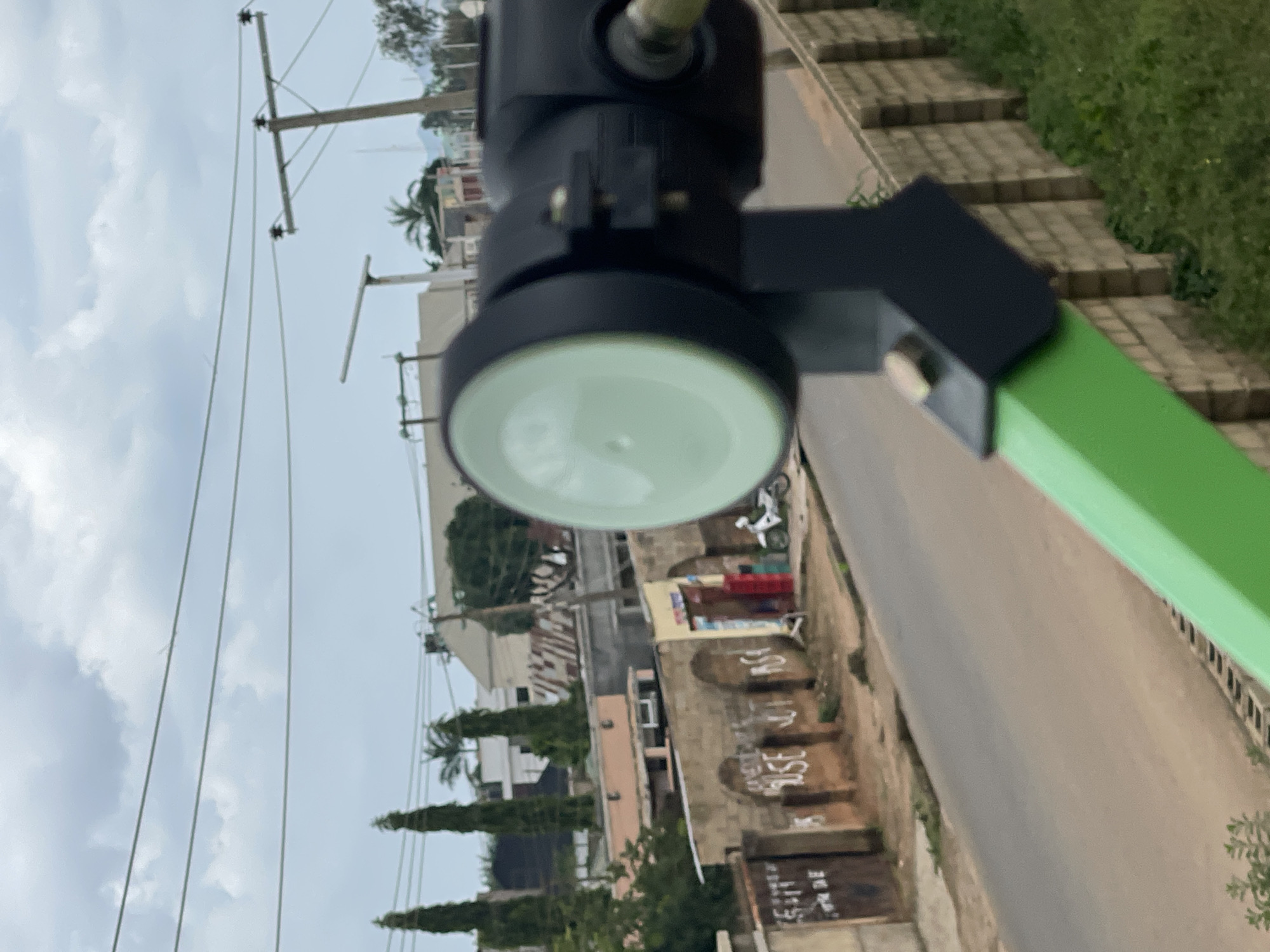 The image shows a close-up of a satellite dish LNB. The LNB is mounted on a green pole, and the dish itself is partially visible. In the background, there is a street with some buildings and vegetation. A motorcycle is parked on the side of the street. The sky is cloudy, suggesting it might be late afternoon or overcast. The setting appears to be an outdoor environment, possibly a residential or commercial area in Jos, Nigeria, given the provided location context. There is no discernible activity or event taking place, as the image is a still shot focused on the satellite equipment. No specific text is clearly visible in the image, although there are some indistinct markings on the buildings in the background.