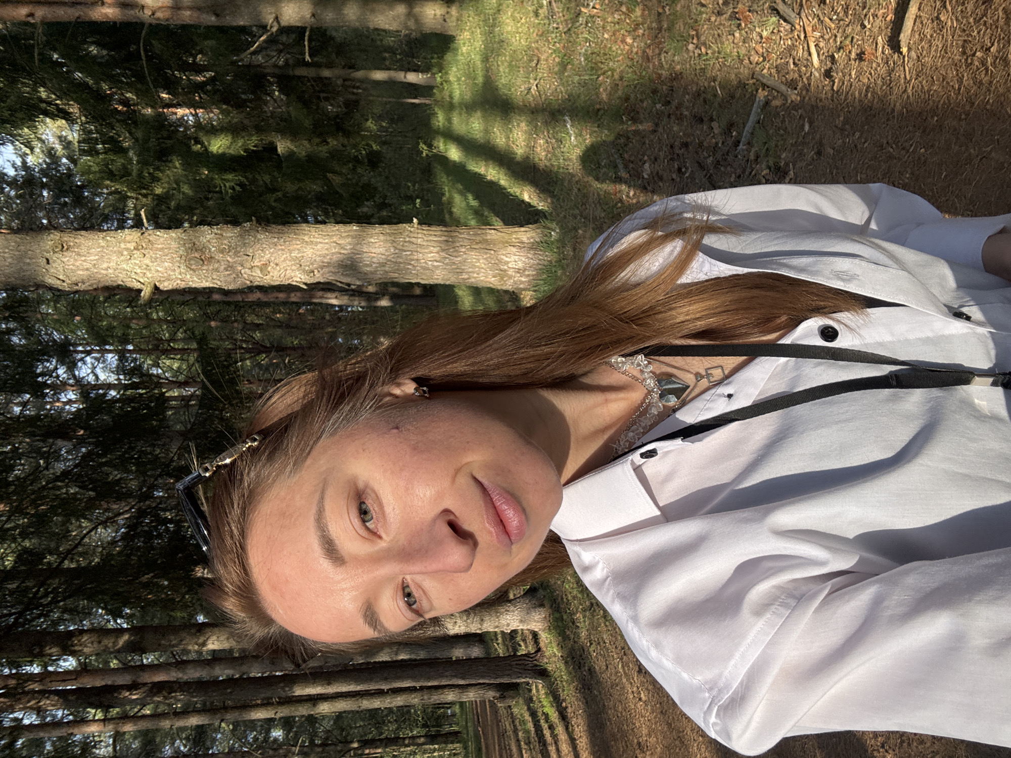 A person is photographed in a forest environment, possibly in Прикамский, Russia. The individual has light skin, brown hair, and is wearing a white button-down shirt. Sunglasses are partially visible on top of their head. The background shows rows of tall trees, suggesting a structured forest or park setting. Sunlight is filtering through the trees, creating areas of light and shadow on the ground and on the person. The photo has been rotated.