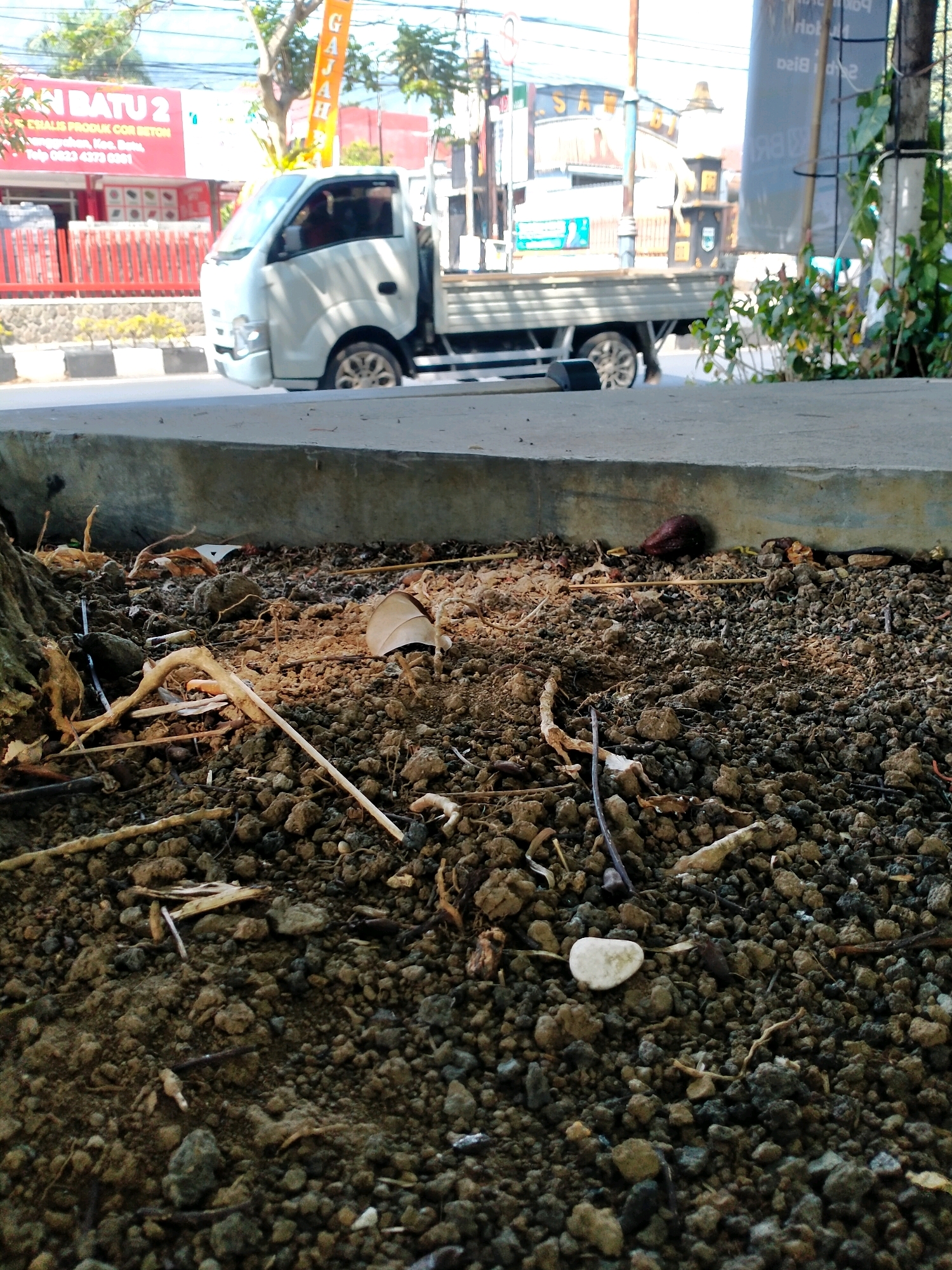 The image captures a close-up view of the ground littered with dirt, small stones, twigs, and fallen leaves. In the background, a white truck is parked on a paved road. To the left of the truck, a building with a red facade and the text "BATU 2" is visible, along with a red metal fence. The scene appears to be located in a city setting, possibly in Batu, Indonesia, given the prominent signage. The lighting suggests it is daytime, with shadows indicating the sun is at an angle.