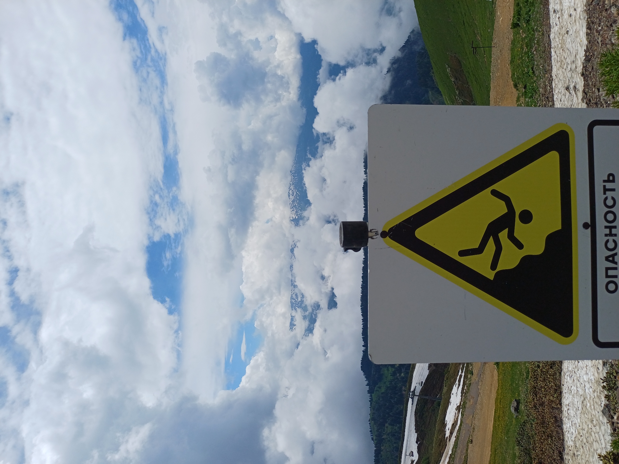 The image shows a warning sign with a falling person pictogram and Russian text that translates to "DANGER" in the foreground. The sign is positioned on a mountainside with a dramatic cloudy sky and distant snow-capped mountains visible in the background. The location is identified as Estosadok, Russia. The environment appears to be a ski resort area, with ski lifts and slopes visible in the middle ground. There are patches of snow and green grass, indicating a transition between seasons or a high-altitude location. The overall scene suggests a potentially hazardous mountain environment.