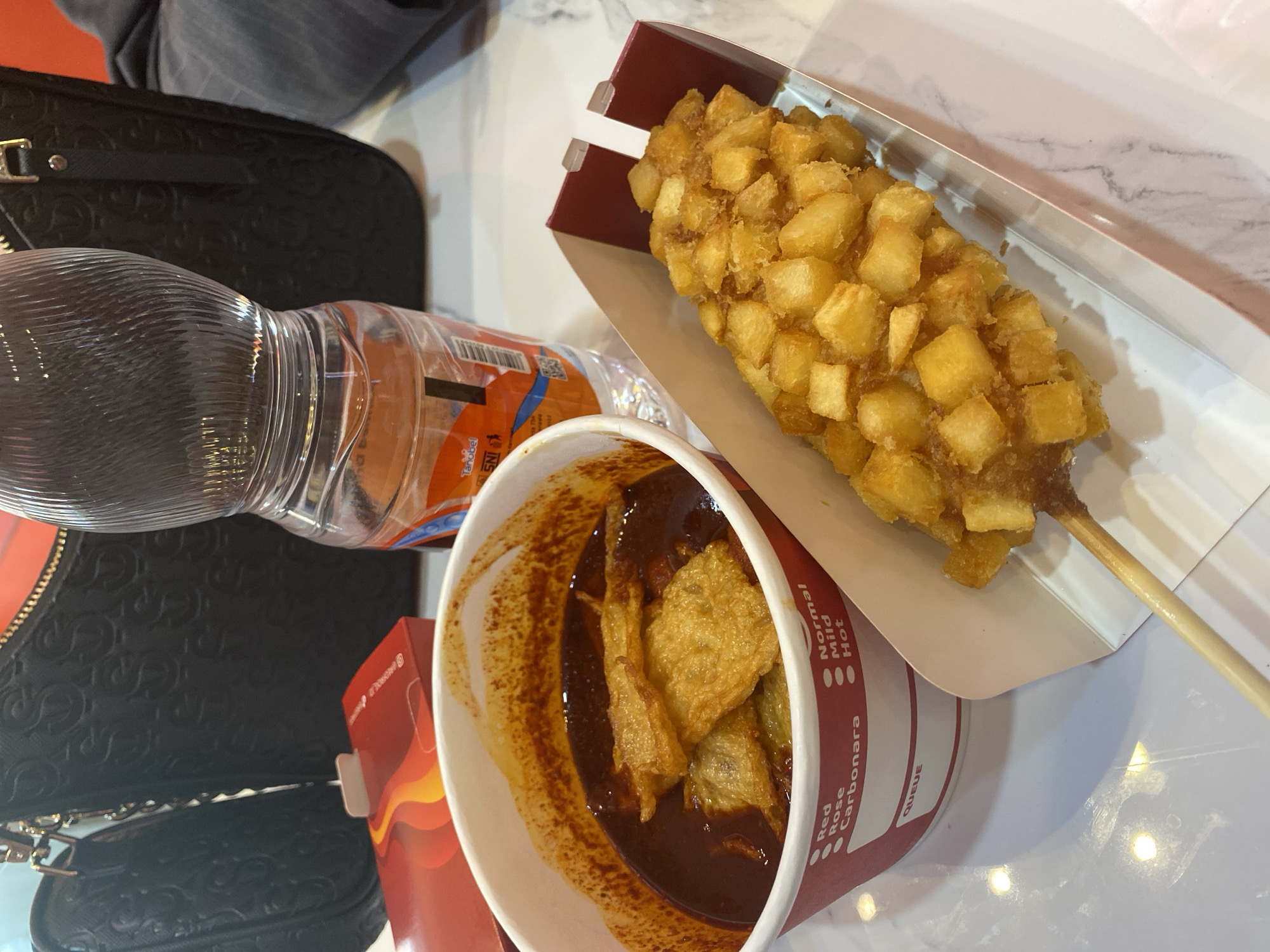 A close-up, eye-level shot captures a meal on a marble countertop, likely at a food establishment in East Jakarta, Indonesia. The focal point is a white cup filled with a dark, thick sauce, possibly tteokbokki, with fried tofu pieces floating on top. To the right, a corn dog coated in diced fried potatoes sits in a red and white cardboard holder, skewered with a wooden stick. In the background, a clear plastic water bottle with a red label is visible, partially obscuring a black patterned handbag with gold hardware. A red box with a flame design lies to the left of the cup. The overall scene suggests a casual dining experience.