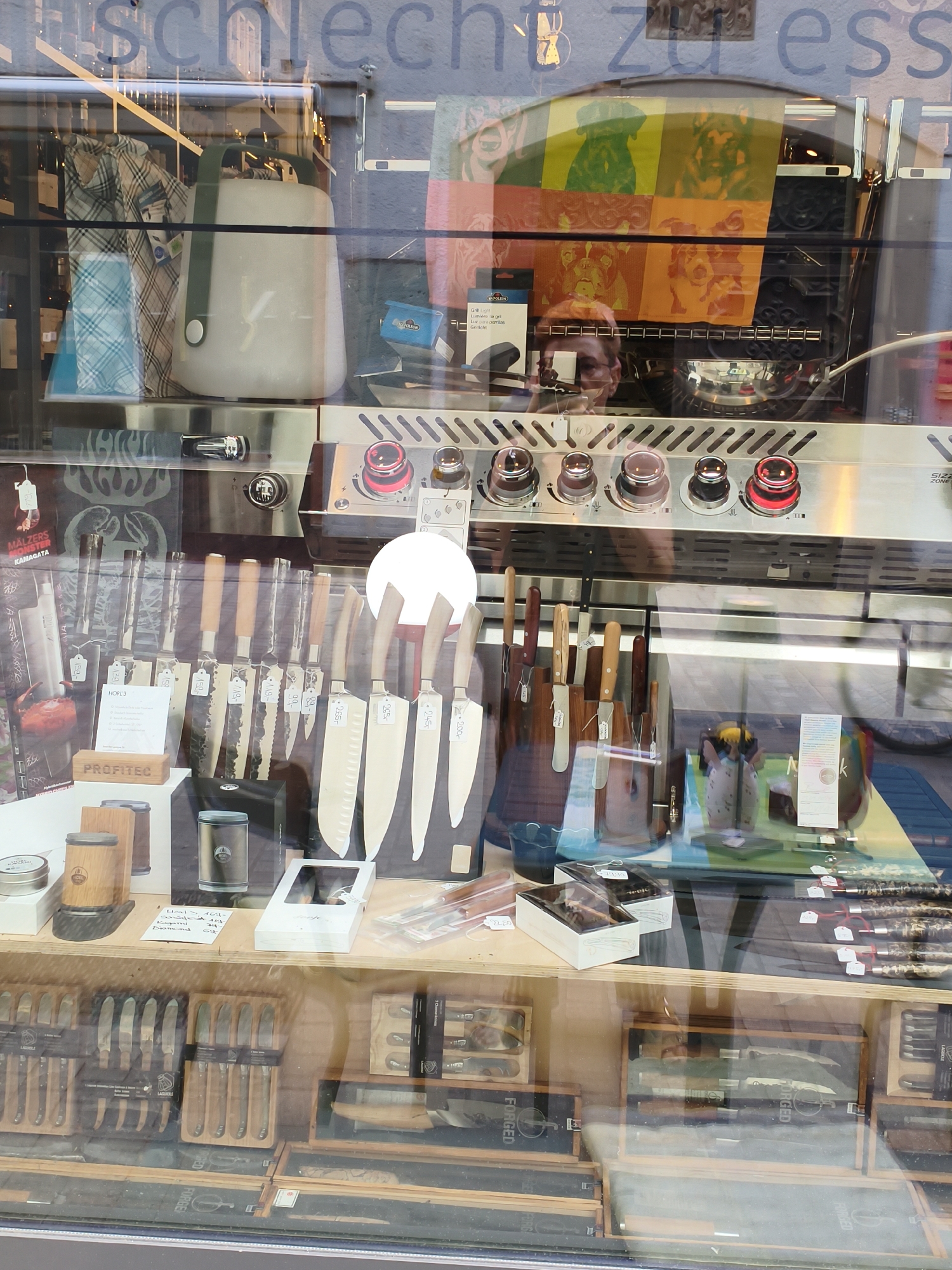 The image shows a display window of a store. The store appears to sell kitchenware, specifically knives, and possibly grills or other cooking appliances.
In the upper portion of the display, a light gray portable lantern with a dark green handle is visible, placed next to what looks like folded fabric or garments. Behind these items, a colorful graphic featuring dog portraits is displayed. A reflection of a person's face is visible in the glass, looking at the display.
Below this, a stainless steel grill with multiple knobs and red illuminated elements is showcased. A row of knives with wooden handles is mounted on a wooden block on a shelf in front of the grill. These knives have price tags attached. To the right of these knives, several larger, sharp-looking kitchen knives with lighter wooden handles are displayed vertically.
On the lower shelves, more knife sets are visible, housed in wooden boxes and blocks. Some of these sets have branding that appears to be "FORGED" and "LAGUIOLE". The overall impression is a retail environment focused on high-quality kitchen tools.
The text visible in the upper part of the window, partially obscured by reflections, appears to be in German and includes "schlecht zu ess" (bad to eat), likely part of a larger advertising slogan. A sign in the middle of the display mentions "PROFITEC". Based on the presence of the German text and the typical retail landscape, this storefront is likely located in the city of Regensburg, Germany.