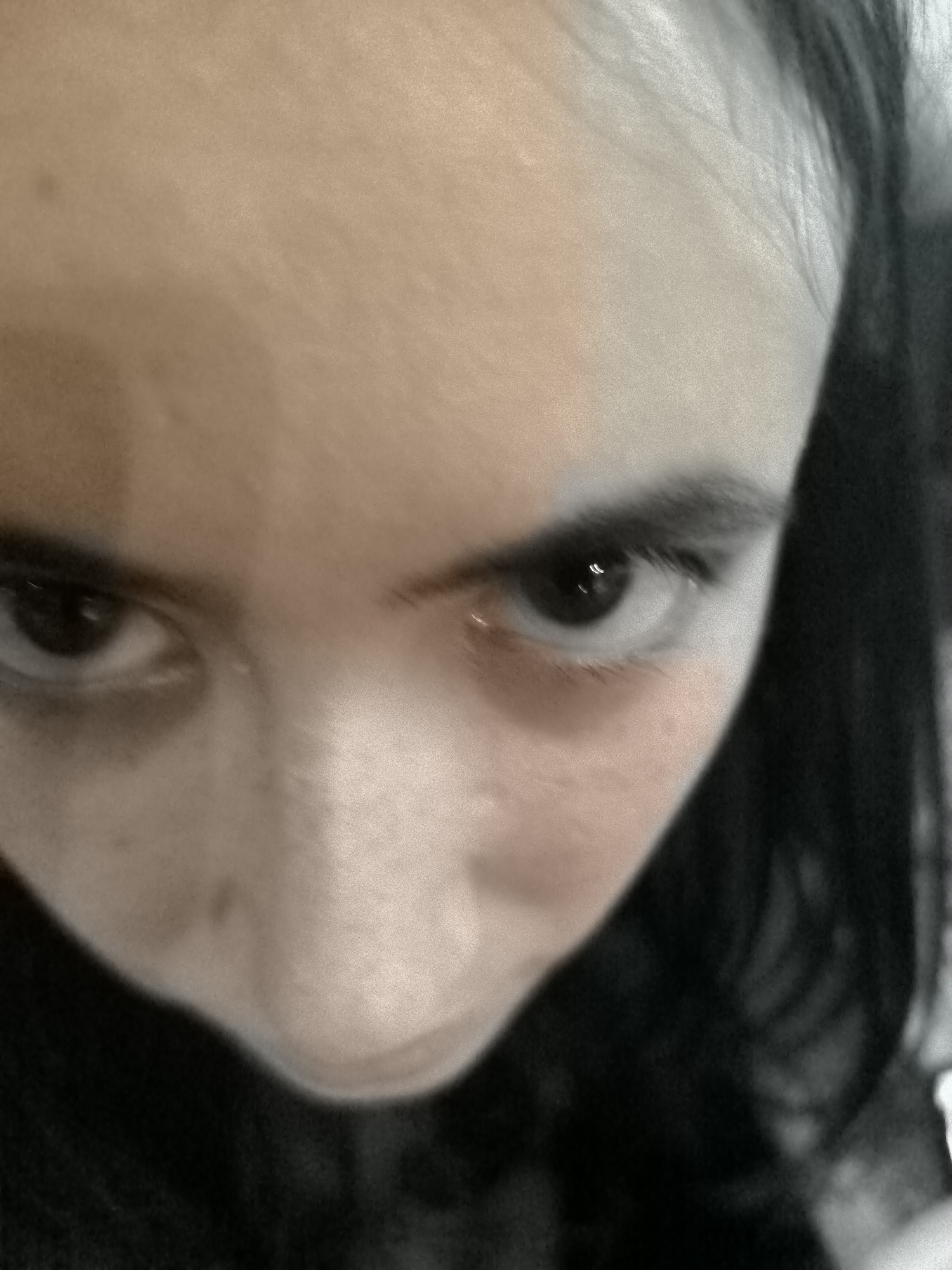 A close-up, low-angle, distorted photographic portrait of a young person's face, focusing on their eyes, is presented. The subject's gaze is directed towards the camera. Their left eye is clearly visible, with a dark iris and pupil, and subtle highlights reflecting light. The area beneath the left eye shows a reddish tint, possibly indicating redness or bruising. Eyelashes are present. The right eye is partially visible and also shows a dark iris. The skin tone is fair with a pinkish hue on the cheeks. The background is dark and out of focus, with strands of dark hair visible on the right side of the frame. The overall image has a grainy, sepia-toned quality. The image does not contain any identifiable landmarks or elements that would allow for its specific location within Cetinje, Montenegro to be determined.