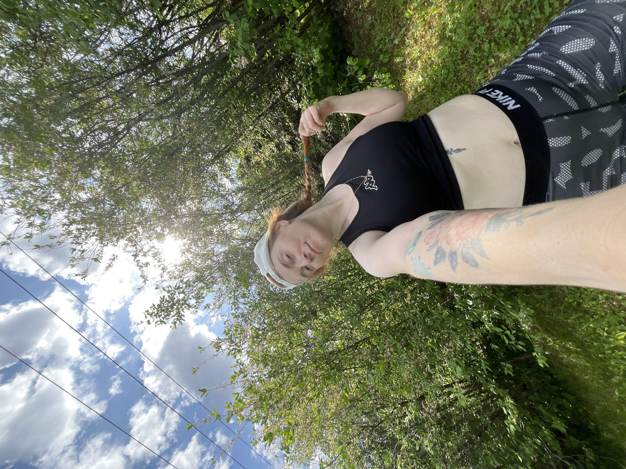 A woman is lying on grass outdoors, appearing to take a selfie. She is wearing a black tank top, patterned leggings with a black Nike waistband, and a light-colored bandana. The sky with clouds and what appears to be power lines are visible. Trees and foliage surround the woman. The location cannot be determined as Chaikovsky, Russia without additional information.