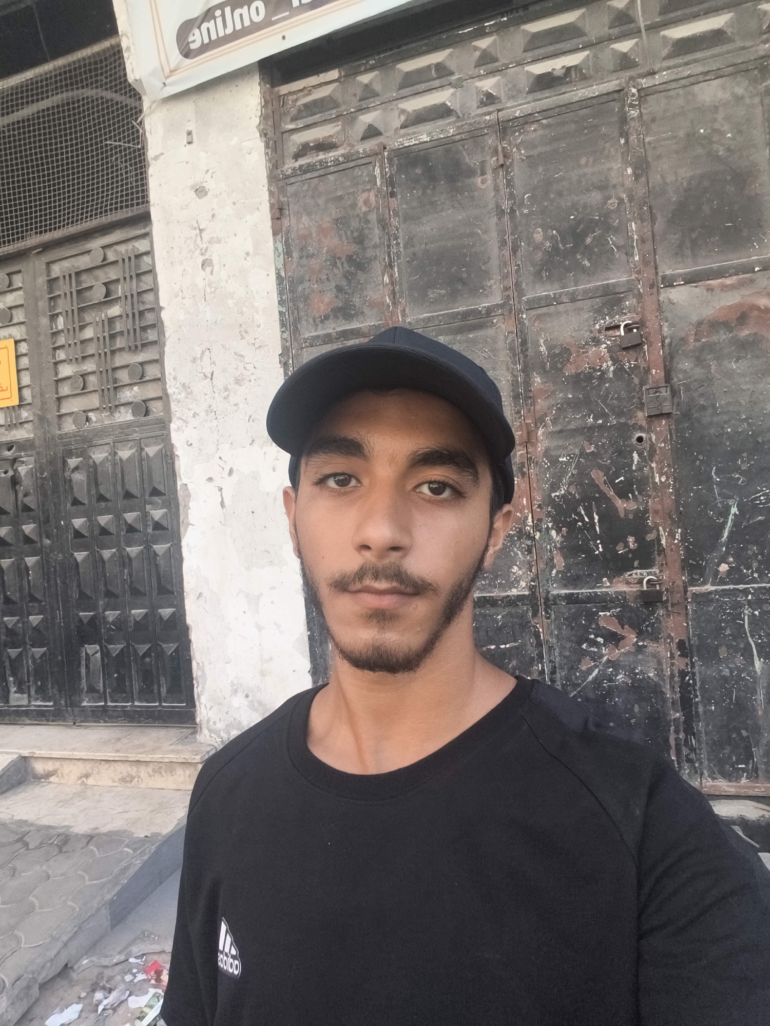 In this medium shot, a young man with a beard and a black baseball cap poses for a selfie in front of a weathered, dark metal door. He is wearing a black t-shirt with a white Adidas logo on the left chest. The background features an old, textured wall with peeling paint to the left of the door and what appears to be a grimy, multi-paneled metal door with visible rust and scratches. The door on the left has a metal grate overlay and a slightly lighter, crumbling wall section. The ground in front of the entrance is paved with gray stones. The overall setting suggests an urban environment in Gaza, Palestinian Territories, characterized by aged architecture and a sense of decay.