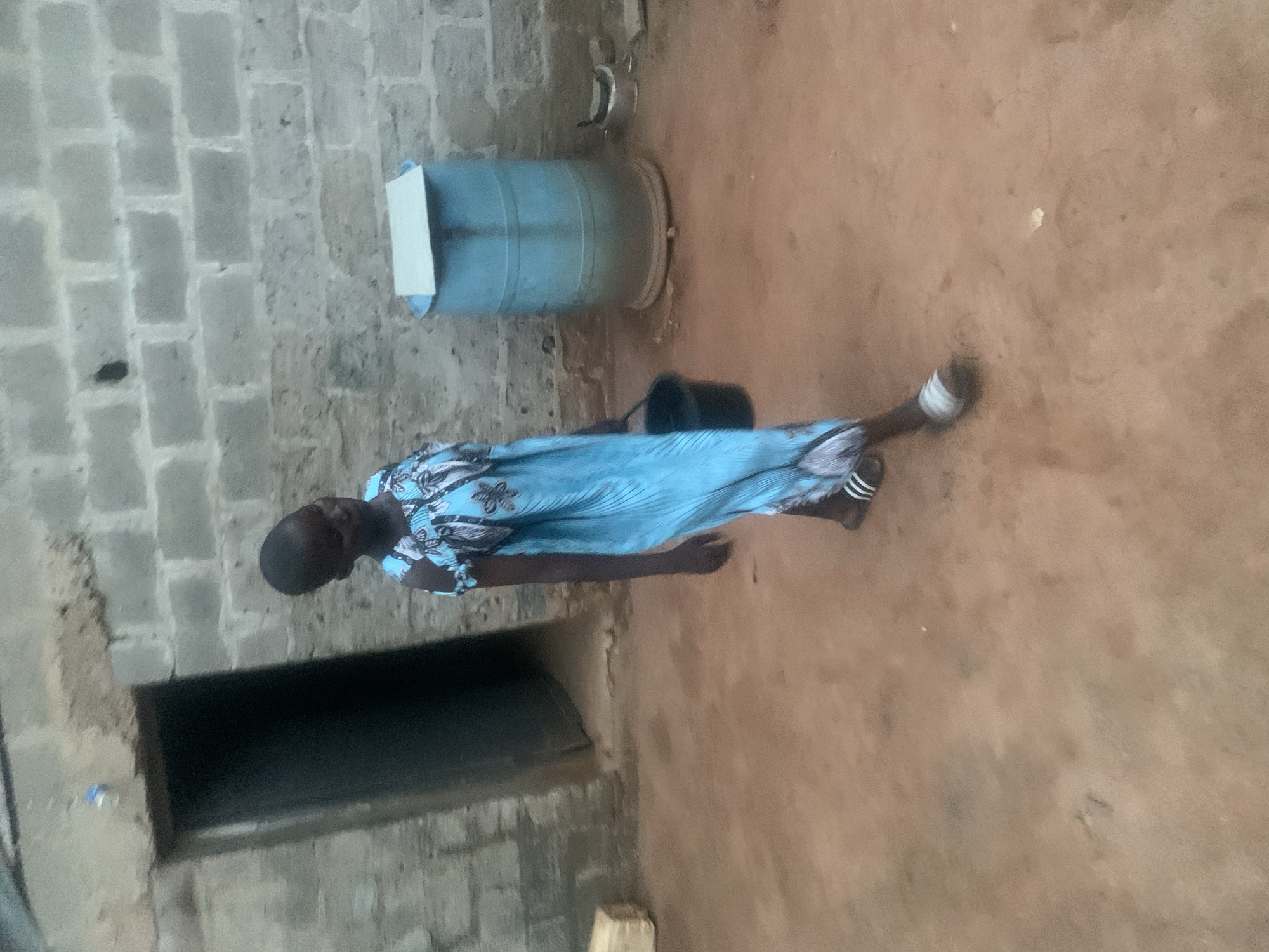 The media file shows a person standing in an outdoor area with a brick wall in the background and a dirt or earthen ground. The person is wearing a blue dress with a floral pattern. They are also wearing sandals with white stripes. The person's left leg is bent at the knee, and their right leg is extended. In the background, there is a large blue barrel with a white lid. To the left of the barrel, there is a dark opening, possibly a doorway or window, set into a rougher wall structure. The lighting suggests it is daytime, possibly late afternoon or early morning given the shadows. The overall scene appears to be in a modest, possibly rural or semi-urban setting. There is no visible text in the image.