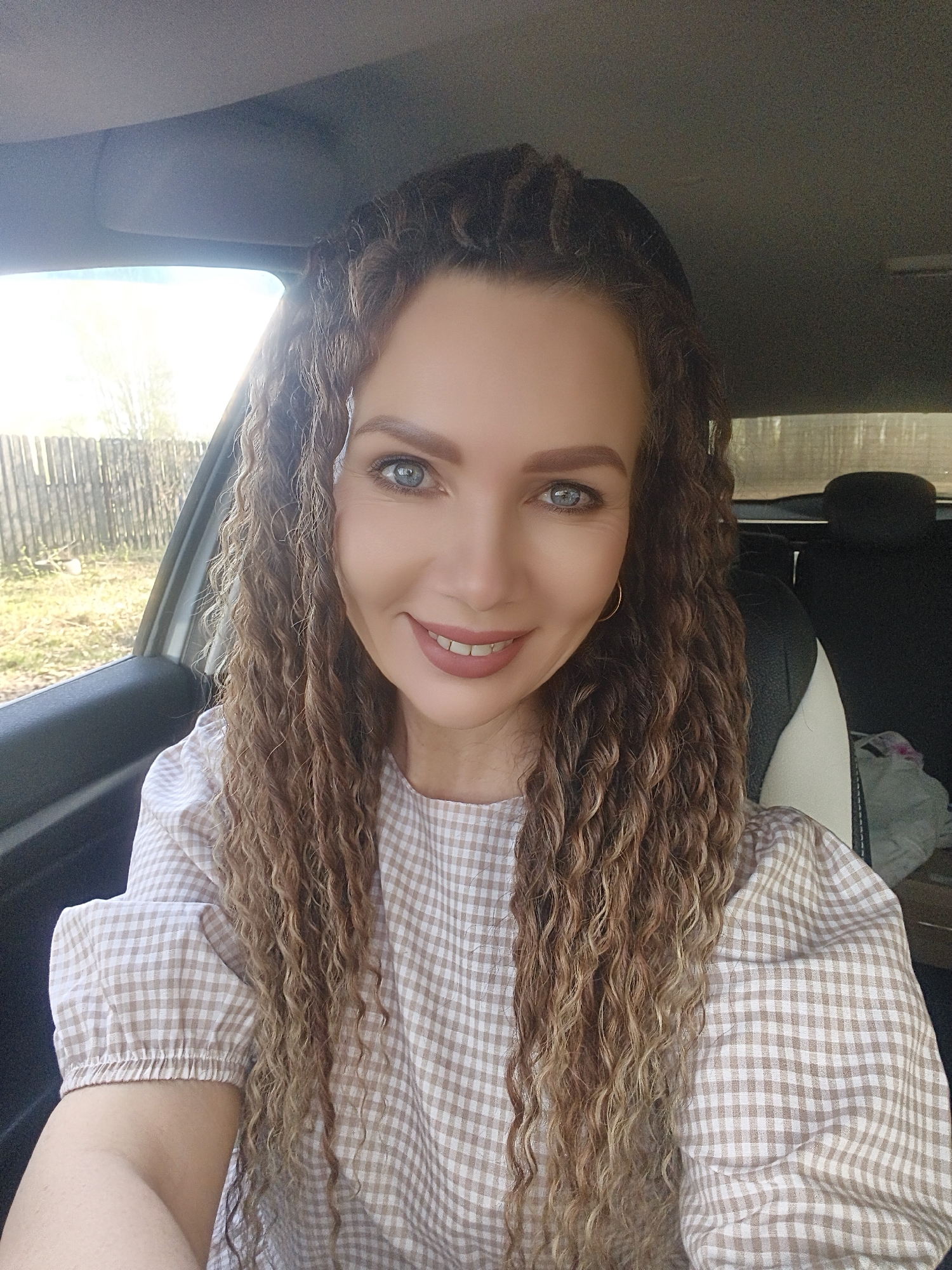 A woman with curly brown hair is taking a selfie inside a car. She is wearing a beige and white checkered shirt and gold hoop earrings. The background shows a car window and a fence outside. The city is Unknown, Unknown.