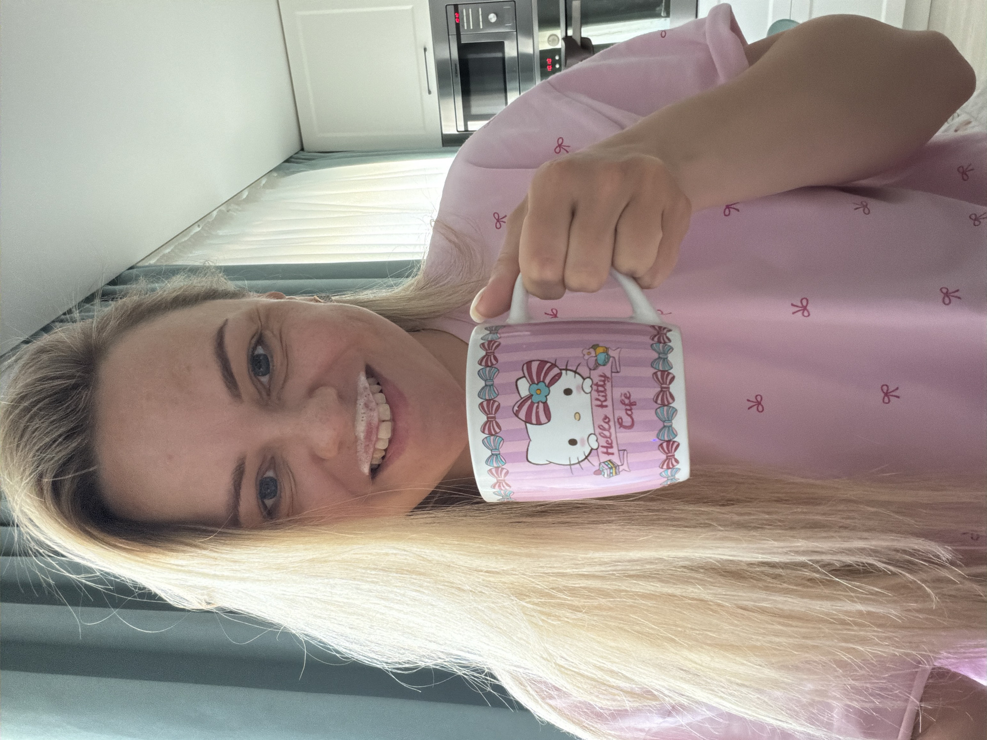 A woman with fair skin, blonde hair, and blue eyes is holding a "Hello Kitty Cafe" mug with pink stripes and bows. She has a milk mustache and is wearing a pink shirt with bow patterns. The background includes white cabinets, a built-in oven, and blinds. There is no indication of Votkinsk, Russia.