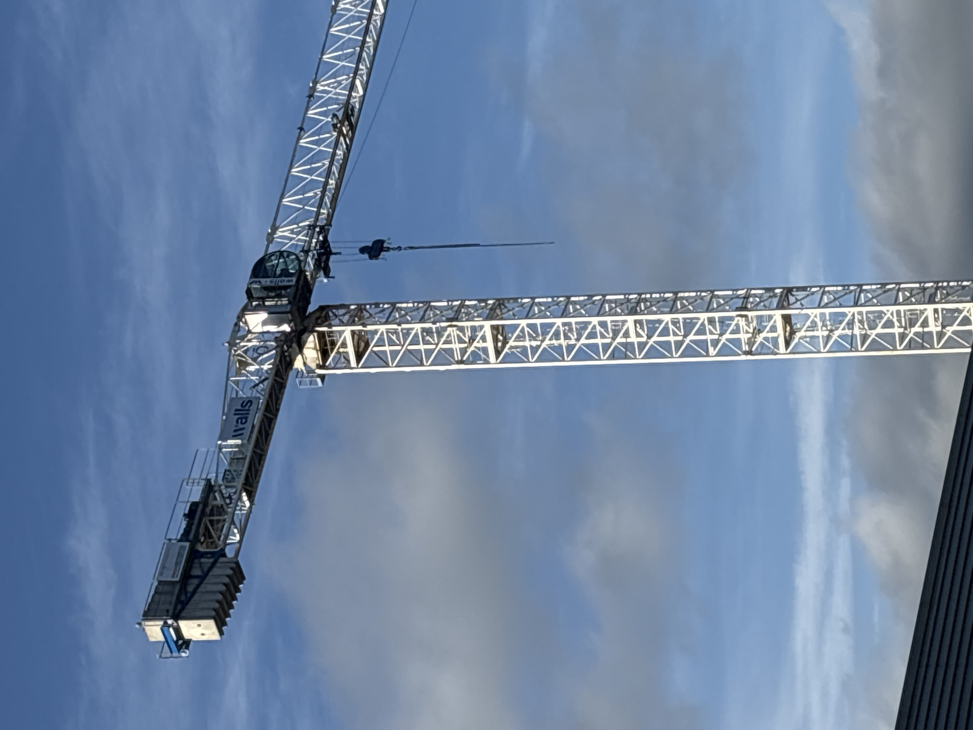An image shows a construction crane against a blue sky with scattered clouds. The crane is white and has the word "walls" visible on its jib. The jib extends from the left side of the image towards the right. A dark, sloped roofline is visible in the bottom left corner of the image. The image does not contain any information about a specific location in the city of Cork, Ireland.