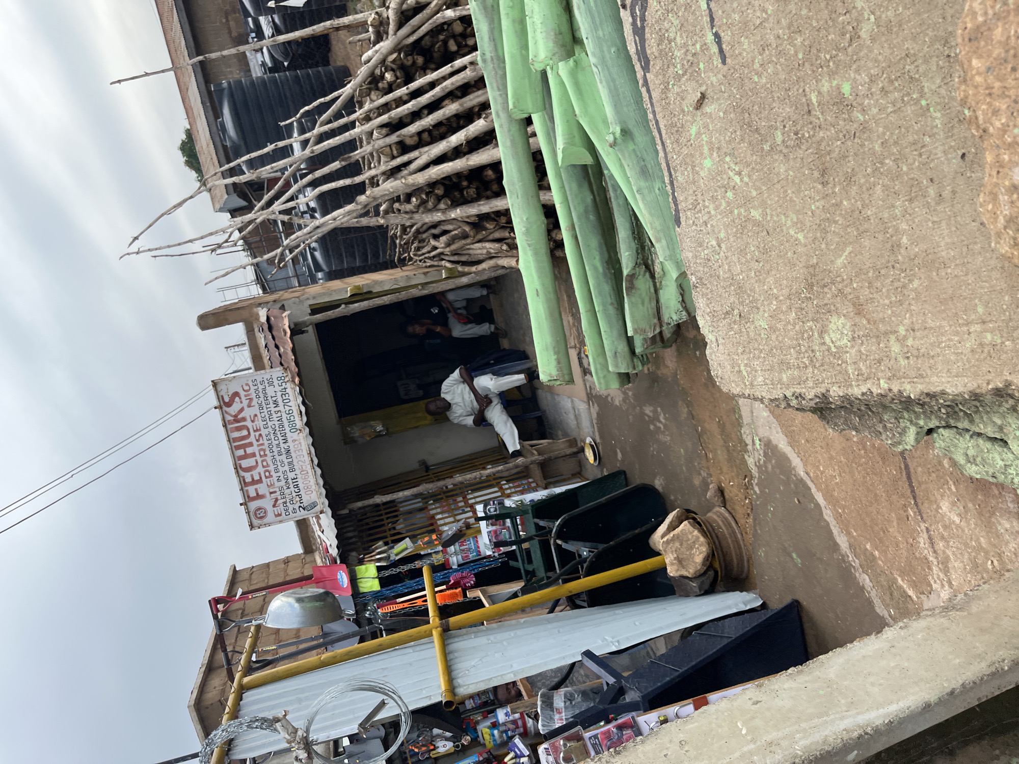 The image depicts an outdoor scene in Gura Top, Nigeria, likely a marketplace or commercial area. In the foreground, a variety of building materials and tools are displayed. These include large, pale green pipes, stacks of dried wood or poles, wire fencing, and metal sheeting. A red shovel and a large metal bowl are also visible. In the background, the facade of a shop is partially obscured by the materials. A sign above the shop entrance reads "FECHUK'S ENTERPRISES NIG. DEALERS IN BUSH-POLES, ELECTRIC POLES & ALL KINDS OF BUILDING MATERIALS. 2nd GATE, BUILDING MATERIALS MKT., JOS. -08060523399 08156703458." Inside the shop, at least two people are present, one appearing to be seated. The overall impression is one of a bustling commercial establishment selling construction-related items. The sky is overcast, suggesting a cloudy day. The ground is uneven and appears to be a mix of concrete and dirt. The image is oriented with the top of the scene to the left.