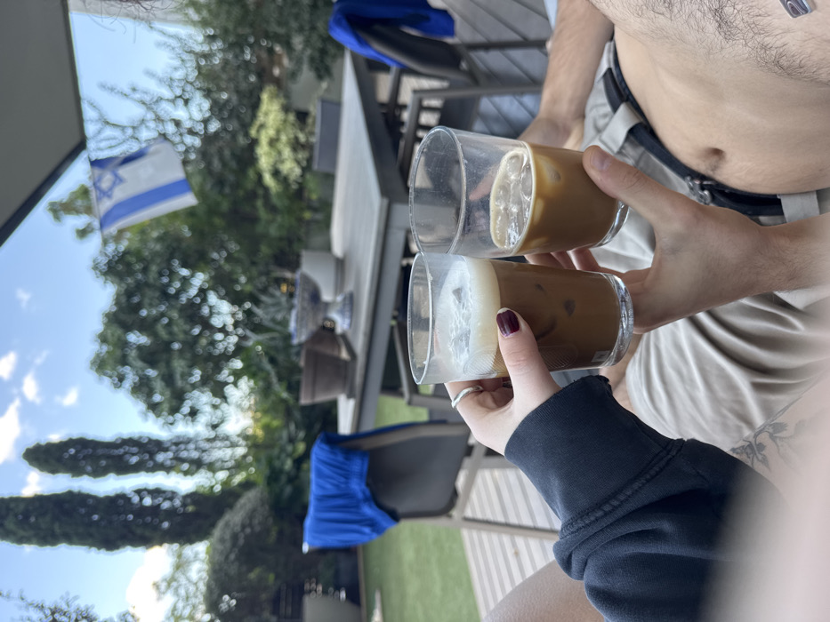 The image depicts an outdoor residential patio and garden scene in Ramat HaSharon, Israel, under a daytime sky with scattered clouds. Two individuals are partially visible from a low angle, holding clear glasses of iced coffee. The person on the left presents a hand with dark red manicured nails and a silver ring on the middle finger, wearing a dark long-sleeved garment and light-colored shorts. This hand holds a glass containing iced coffee topped with a thick layer of white foam. The person on the right displays a bare, hairy abdomen and a hand, wearing light-colored shorts with a visible dark belt. This hand holds a glass of iced coffee with visible ice cubes and less foam. The glasses are held together, indicating a toast or clinking action.
In the background, a light-colored rectangular outdoor dining table is visible, adorned with several potted plants and a blue and white decorative ceramic bowl. A blue towel or piece of clothing is draped over a dark-colored chair situated to the left of the table. Beyond the patio, a lush green garden extends with various trees and shrubs, including two tall, slender cypress-like trees on the left. An Israeli flag, featuring blue stripes and a Star of David on a white background, is partially visible, unfurled against the green foliage in the upper right portion of the frame. A grey awning or roof structure is visible at the very top. The ground beneath the furniture consists of both green lawn and a section of light-colored wooden decking.