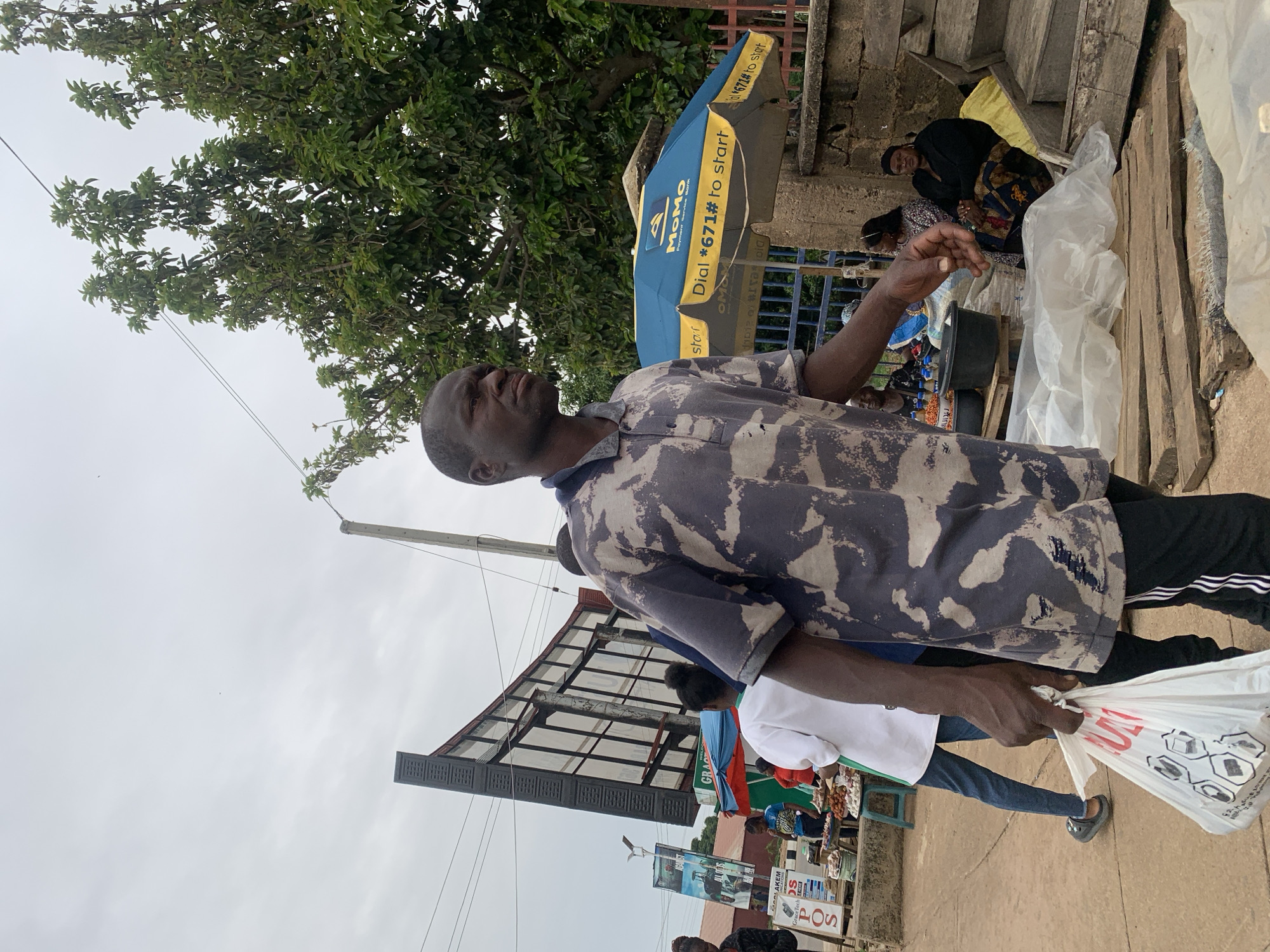In Jos, Nigeria, a man walks down a street, carrying a white plastic bag in his right hand. He wears a short-sleeved, camouflage-patterned polo shirt and dark pants with white stripes. The man's gaze is directed to his right, and he appears to be walking through a busy outdoor market. In the background, an open umbrella, branded "MoMo Payment Service Bank," displays the text "Dial *671# to start." Several market stalls are visible, with goods displayed on tables. A large billboard is positioned behind some of the stalls. Trees with green foliage line the right side of the frame, and a cloudy sky stretches overhead. The ground is paved with what appears to be concrete or asphalt, with some wooden planks scattered in the foreground. Other people are present in the background, some appearing to be vendors and others shoppers.