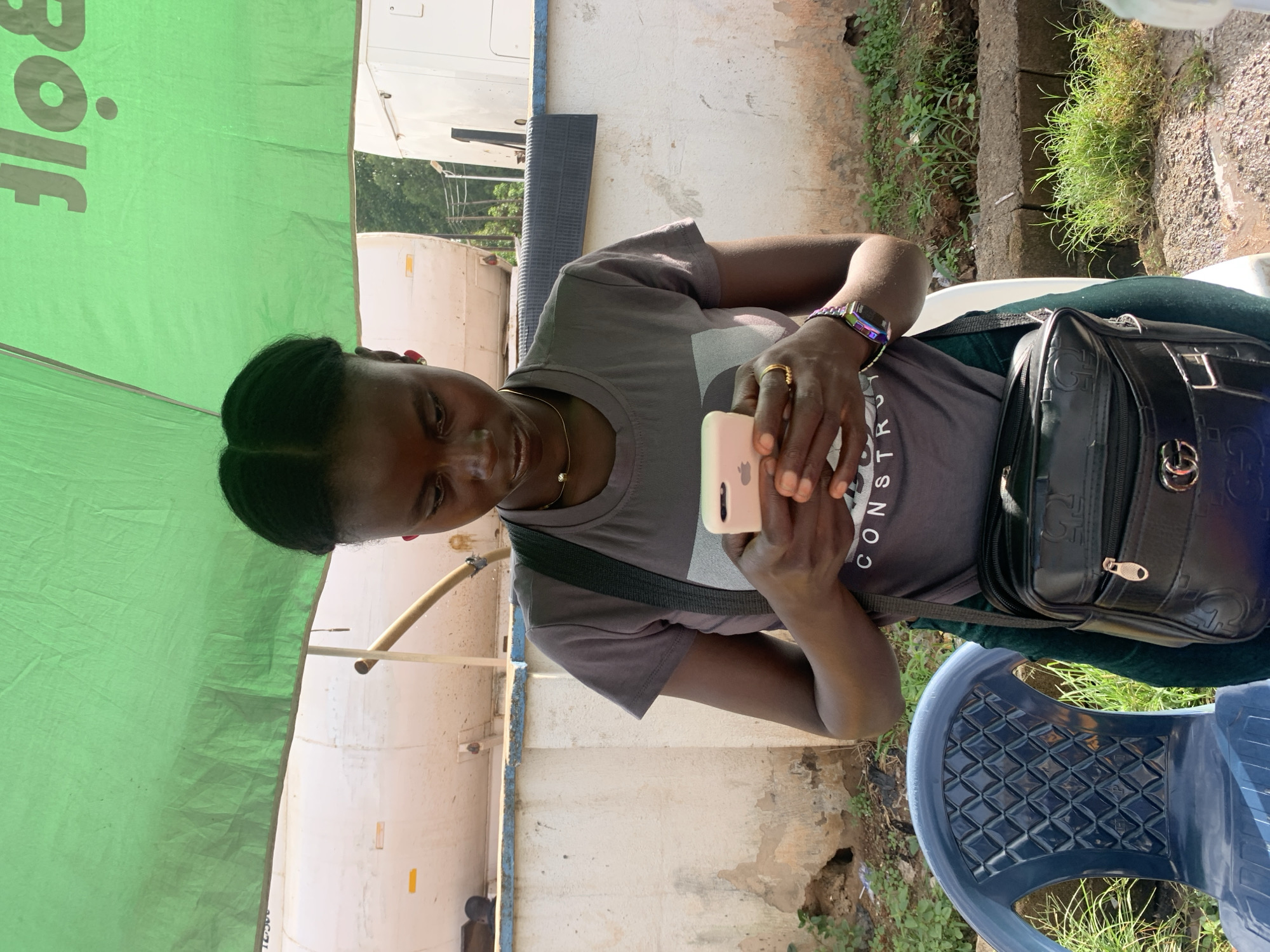 A young Black woman, with dark braided hair and wearing a gray t-shirt, is standing outdoors, looking down at her pink smartphone. She is wearing a thin gold necklace and a ring on her right ring finger. A smartwatch with a colorful band is visible on her left wrist. A dark crossbody bag with a gold zipper and embossed pattern rests on her right hip. She is positioned in front of a white concrete wall, with a portion of a blue plastic chair to her left. Behind her, a large green canopy provides shade, and glimpses of white tanker trucks and trees are visible. The ground is a mixture of dirt and sparse green vegetation. The scene appears to be in a dusty outdoor area, possibly a marketplace or roadside.