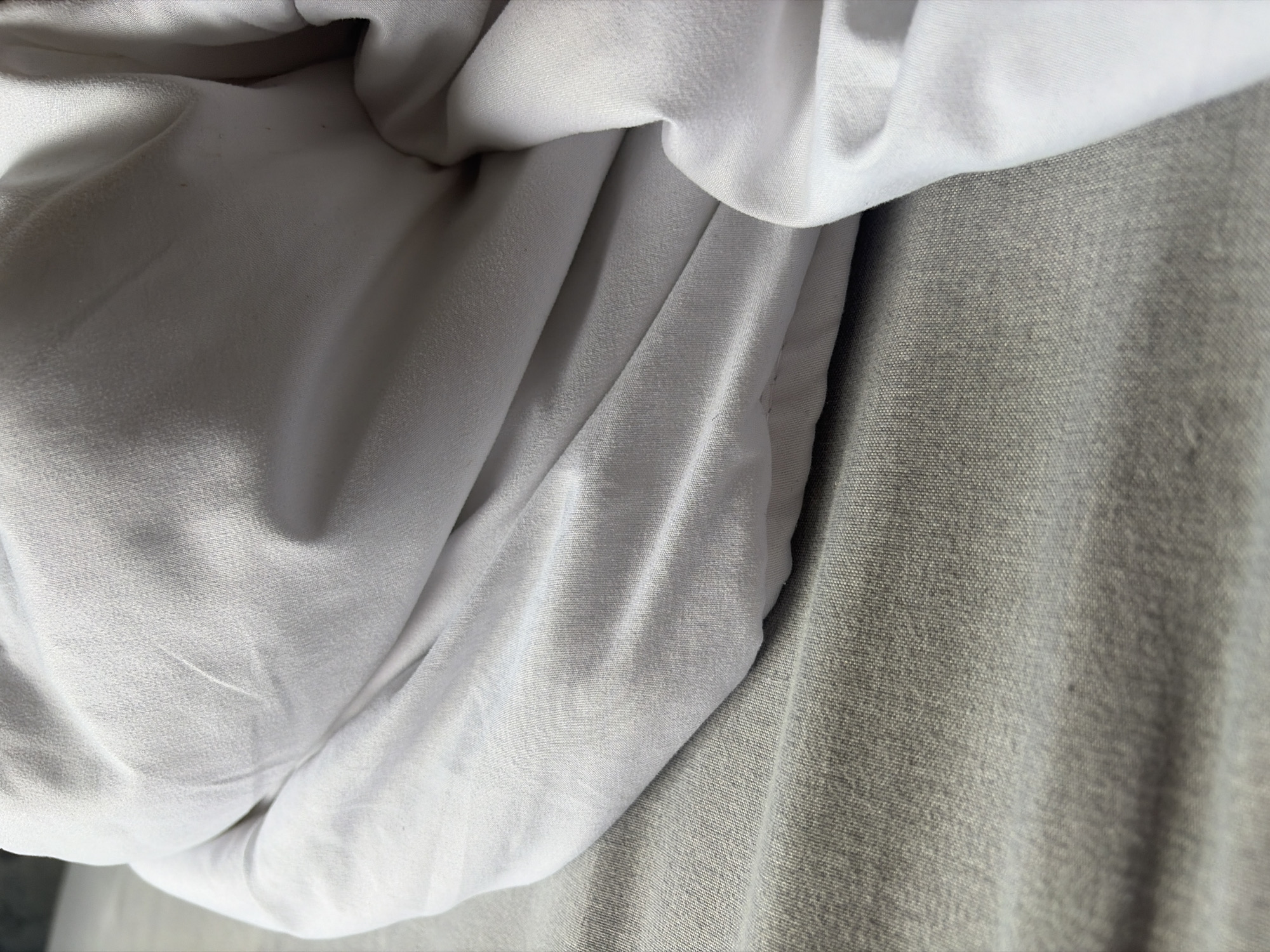 A close-up, overhead shot of a white duvet draped over a grey textured surface, possibly a bed or sofa. The duvet is wrinkled and folded, creating shadows and highlights across its surface. The grey surface has a visible woven texture. The image appears to be taken indoors in a neutral setting, with no specific city or location details evident.