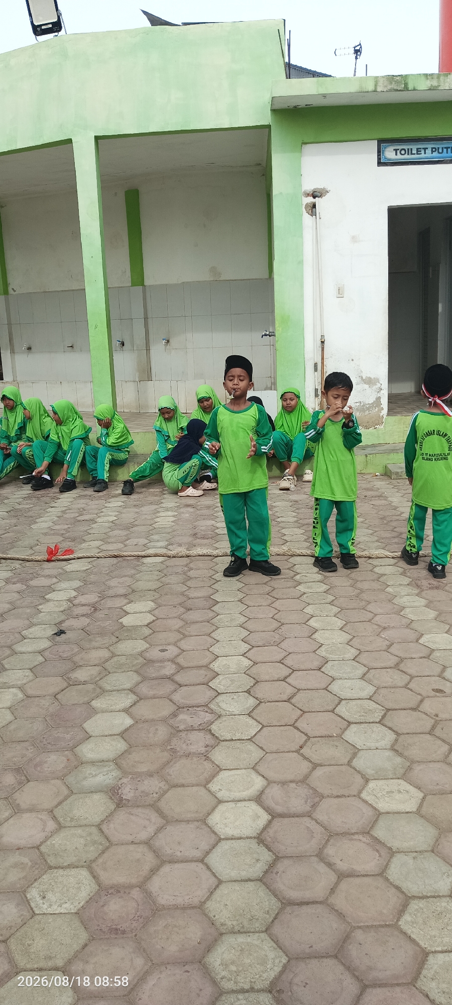 In Blangkrueng, Indonesia, a group of young children wearing matching green hijabs and pants are gathered outdoors. Some are sitting on a tiered concrete structure, while others stand in front of them. Two boys in the foreground, also in green uniforms, are facing the camera and appear to be speaking or singing. A rope lies on the hexagonal-patterned pavement in front of the children. Behind the group, a light green building with a white wall and a sign that reads "TOILET PUTIH" is visible. The scene appears to be a school or community event.
