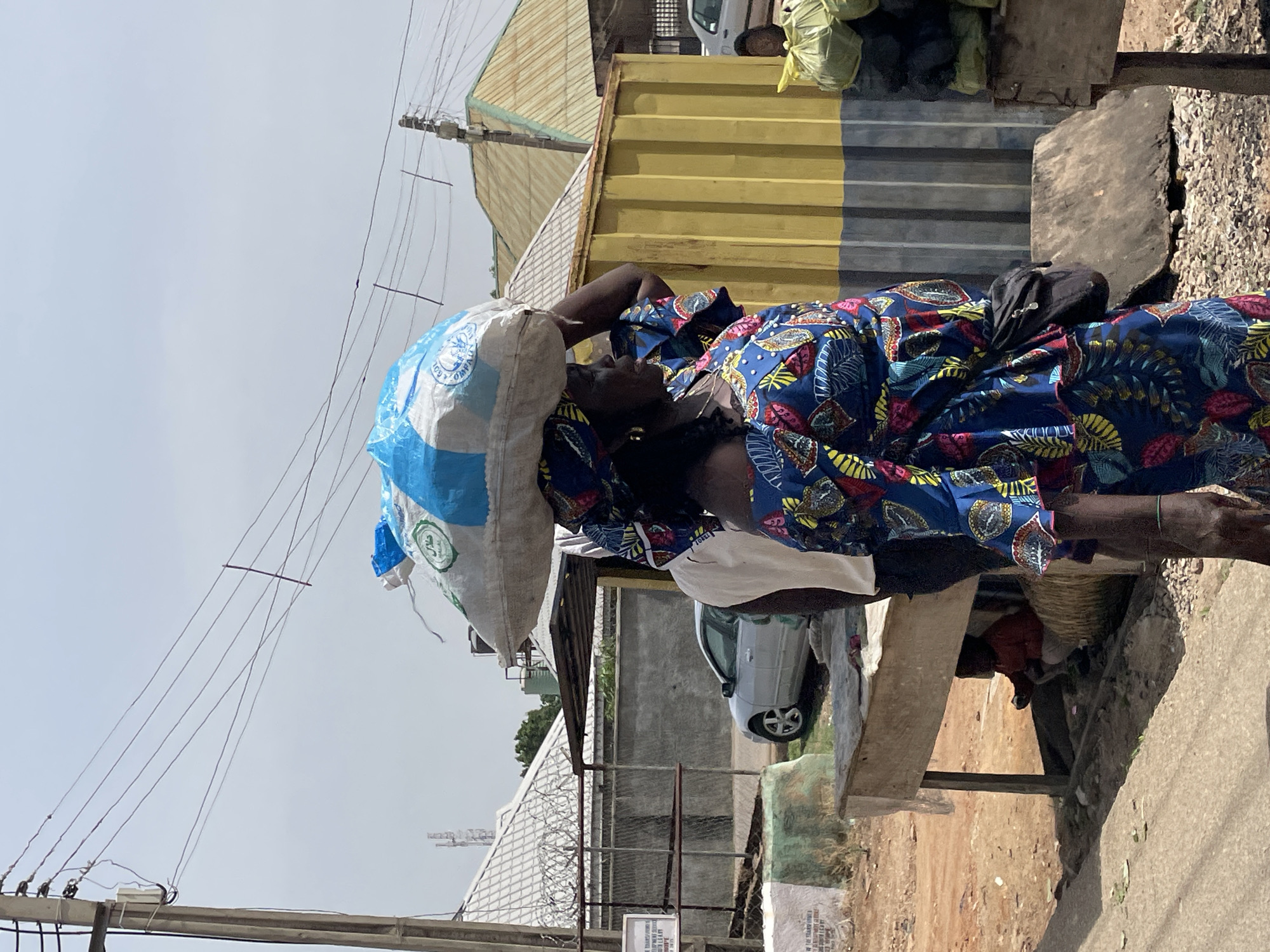 A woman in Jos, Nigeria, balances a large white and blue bag on her head while standing near a vendor's stall. She is wearing a blue, red, and yellow patterned garment. A black bag is resting on her back. A partially visible vendor sits beneath a wooden shelf. Yellow and gray panels and a white car are visible in the background, along with a gray fence topped with barbed wire. Overhead electric wires crisscross the sky.