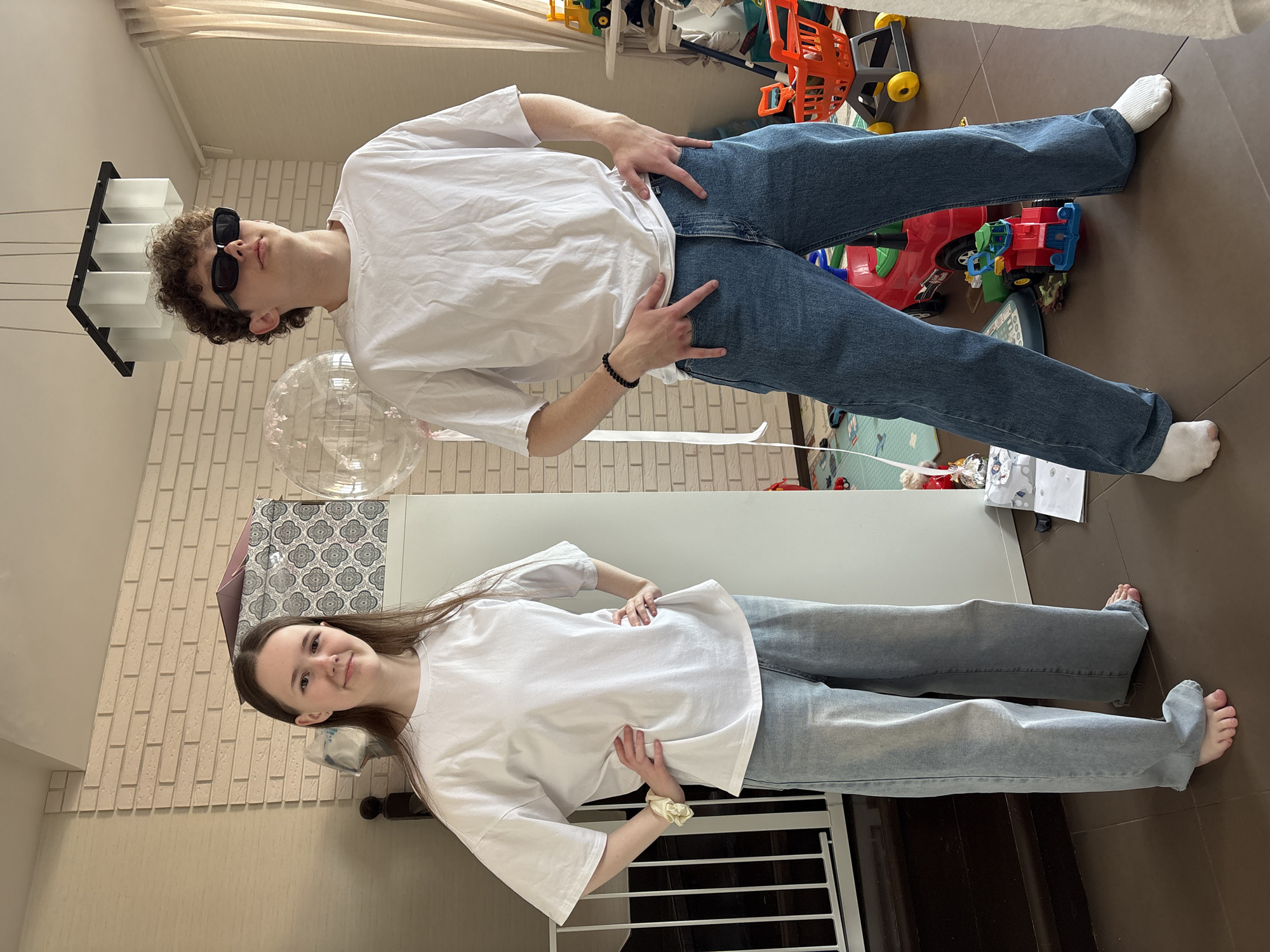 Two individuals, a male and a female, are posing indoors for a photo. The male, positioned in the upper part of the frame, has curly hair and wears sunglasses, a white t-shirt, blue jeans, and a black bracelet. He stands with his hands on his hips, striking a pose. The female, located in the lower portion, wears a white t-shirt and blue jeans, with a hair tie on her wrist, and also poses with her hands on her hips. The background includes a partially visible brick wall, a white table, various toys scattered on the floor, and other domestic objects. The location appears to be a private residence. The information is not available within the given image to determine the location is in Ulyanovsk, Russia.