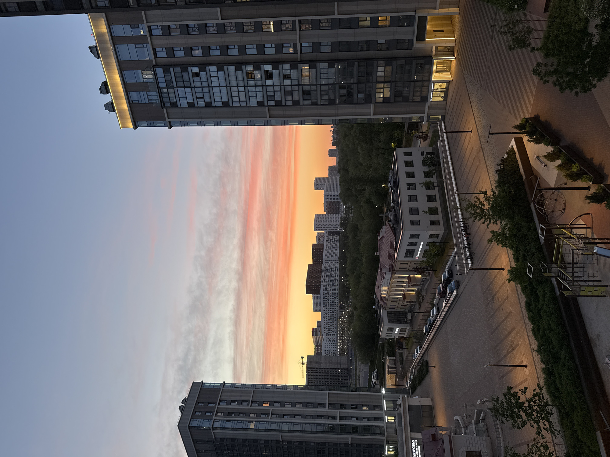Here is a description of the image:
The image depicts an urban scene, likely in Moscow, Russia. The view appears to be from a high vantage point, possibly a balcony or rooftop. Modern, high-rise apartment buildings frame the sky, which displays a sunset or sunrise with orange and pink hues in the clouds. Several buildings are visible in the skyline, some shrouded by trees. A pedestrian walkway with streetlights and decorative shrubbery leads away from the buildings. Cars are parked along the roadside, and other buildings of varying styles line the street.