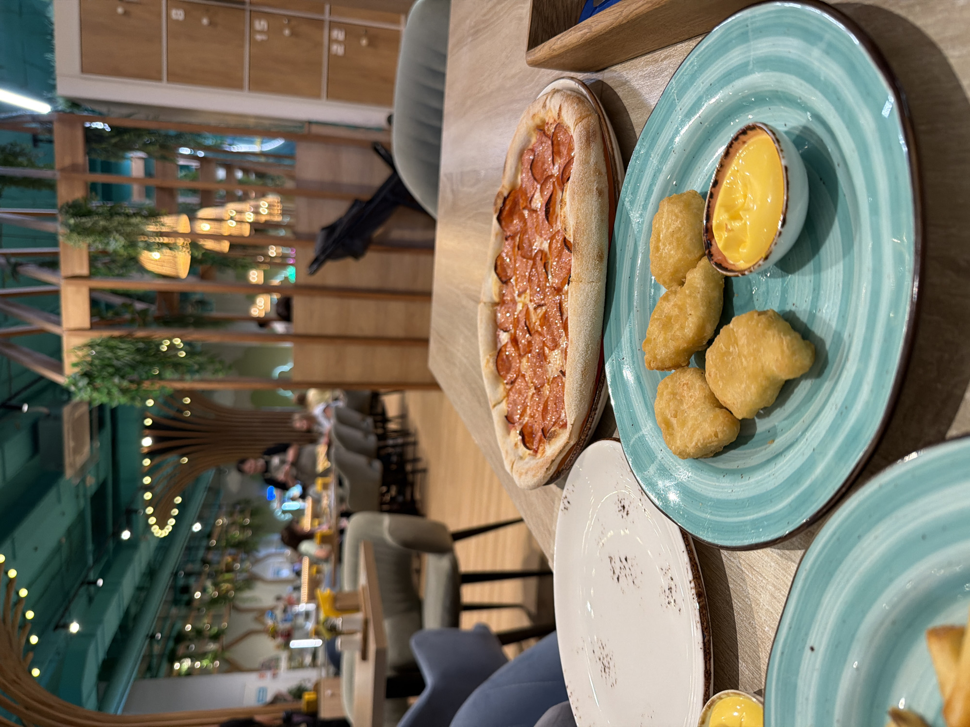 A close-up shot shows a table in a restaurant with food. In the foreground, there is a pizza topped with pepperoni, chicken nuggets with a side of dipping sauce, and an empty plate. In the background, the restaurant's interior is visible, featuring wooden beams, hanging plants, and patrons dining. The restaurant is likely located in Kazan, Russia, given the context.