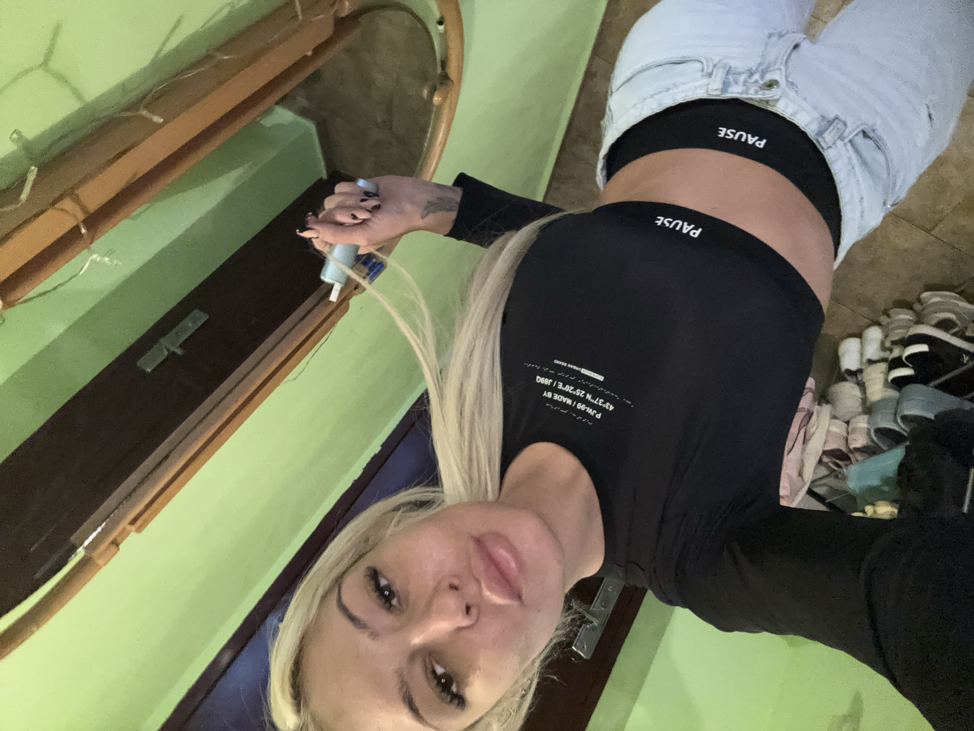 A woman with blonde hair is posing for a selfie. She is wearing a black long-sleeved crop top with the word "PAUSE" on the band, and light-wash jeans with the word "PAUSE" visible on the waistband beneath the jeans. The text "PJW-99/MADE BY 43°37″N 25°20″E / J89Q URBAN BRAND" is printed on the upper left side of her shirt. The coordinates on her shirt, 43°37'N 25°20'E, are not located in Plovdiv, Bulgaria but rather in northern Bulgaria, about 200km away. She is holding a cylindrical object in her right hand. The background includes a green wall, a wooden structure, and a shelf with shoes and other items.
