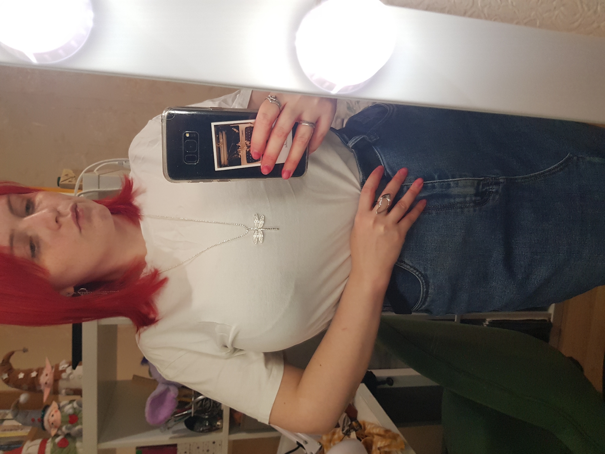 A woman with bright red hair takes a mirror selfie, holding a smartphone. She is wearing a white t-shirt and a denim skirt. The t-shirt has a dragonfly pendant necklace. She has a ring on her right hand. The background shows shelves with various items, including small figurines. A green chair is visible on the left side of the image, and a lighted mirror is on the right.
