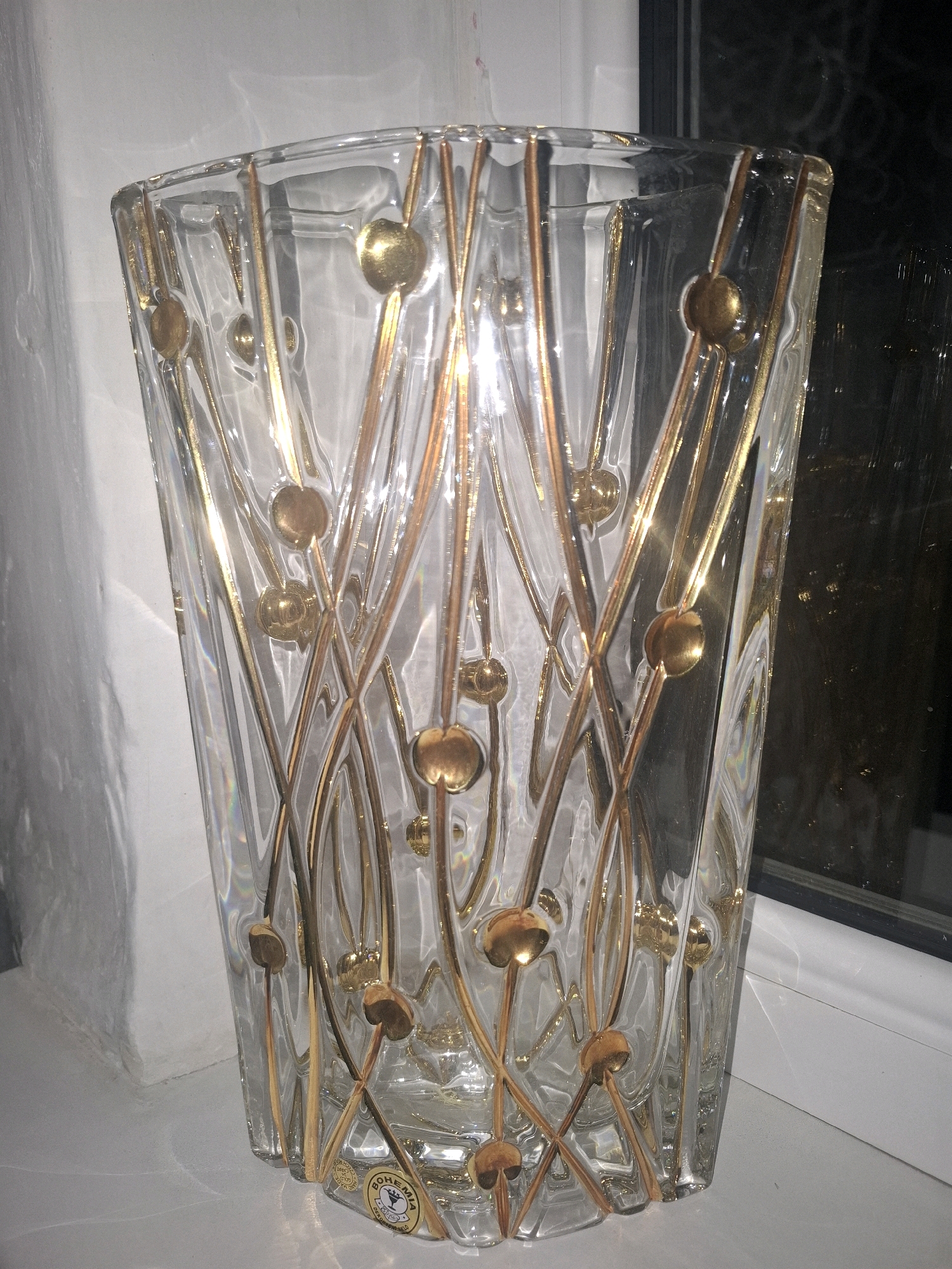 A clear glass vase with gold accents is positioned on a white surface, likely a windowsill, with a blurred background that includes a window with a dark reflection and a textured white wall. The vase is tall and rectangular with a tapered shape, featuring delicate gold lines that weave around the exterior, punctuated by small, round gold spheres. Sunlight reflects off the glass and gold elements, creating bright highlights and subtle rainbow refractions. The bottom of the vase has a circular label with text and a graphic that appears to read "BOHEMIA". The overall scene is well-lit, suggesting it is daytime. The location is Almaty, Kazakhstan.