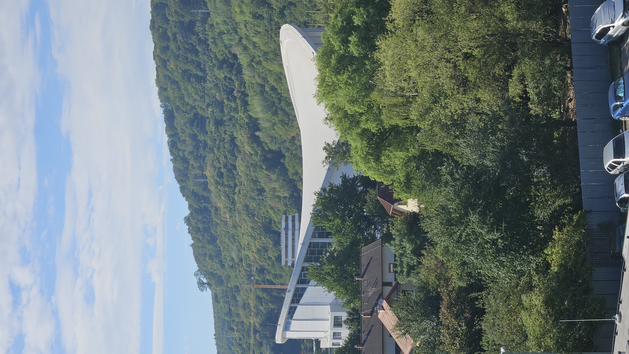 This is an exterior, eye-level, medium shot of a modern building with a curved white roof, situated in the city of Prešov, Slovakia. The building is surrounded by lush green trees and foliage, with a hill covered in trees in the background. In the foreground, several cars are parked in a lot with a corrugated metal fence. The sky above is blue with scattered white clouds. The overall scene depicts a contemporary architectural structure integrated into a natural landscape.