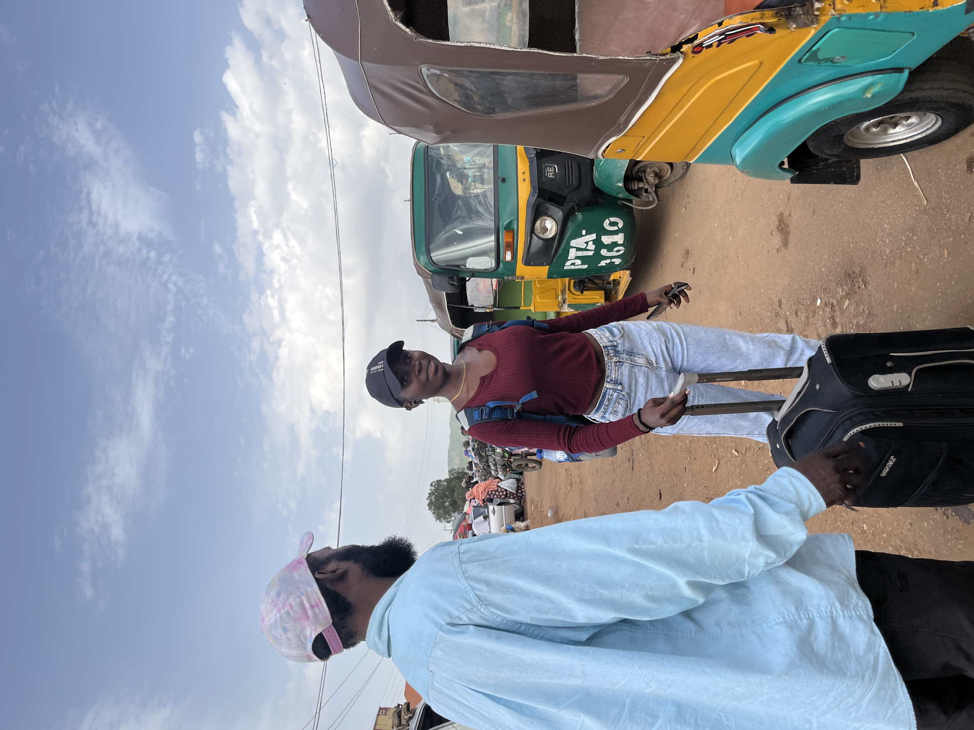 A young woman and a man are standing near a colorful auto-rickshaw, possibly preparing for travel. The woman, wearing a maroon top, light blue jeans, and a black baseball cap, is holding a mobile phone. She has a backpack slung over her shoulder and is standing next to a dark suitcase. The man, in a light blue shirt and a pink and white tie-dye baseball cap, is partially visible in the foreground, also near a suitcase.
The auto-rickshaw is green and yellow with blue accents, and it has "PTA 3610" painted on its front. Other auto-rickshaws and some vehicles are visible in the background, suggesting a transportation hub or busy street. The scene appears to be outdoors, under a bright, partly cloudy sky. The ground is dusty or sandy. The overall impression is a travel preparation scene, possibly in a warm, sunny climate. The people's expressions are neutral, but the context suggests anticipation or readiness for a journey. The time of day appears to be daytime, given the bright sunlight. The location context suggests Jos, Nigeria.