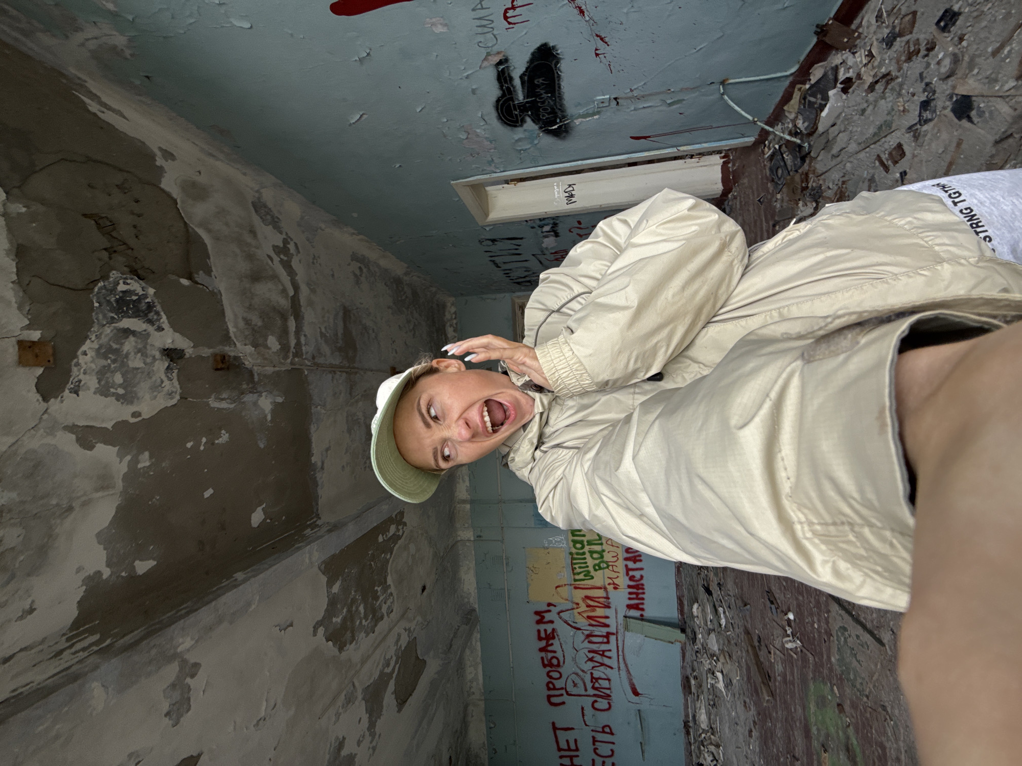 A young woman wearing a light beige jacket and a light green baseball cap is taking a selfie in what appears to be an abandoned building in Teriberka, Russia. The building's interior is in disrepair, with peeling paint on the walls and ceiling, and debris scattered across the floor. The woman has an exaggerated expression of surprise or shock on her face, with her mouth open and her hand near her cheek. Red graffiti is visible on the walls, with some text in Russian. The overall impression is one of urban decay and exploration.