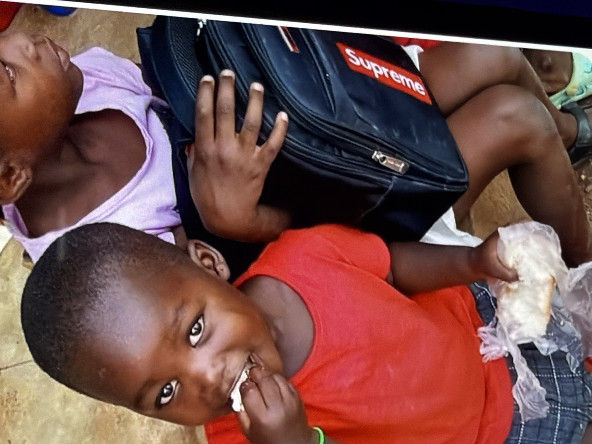 Two young children, one in a red t-shirt and the other in a purple top, are visible in an outdoor setting. The child in the red t-shirt is holding a piece of bread in a plastic bag and appears to be eating. This child is also wearing plaid shorts and a green bracelet on their left wrist. The other child is seated and holding a black backpack with a red "Supreme" logo. The ground beneath them appears to be a dirt or stone surface. The scene is a close-up shot of the children.