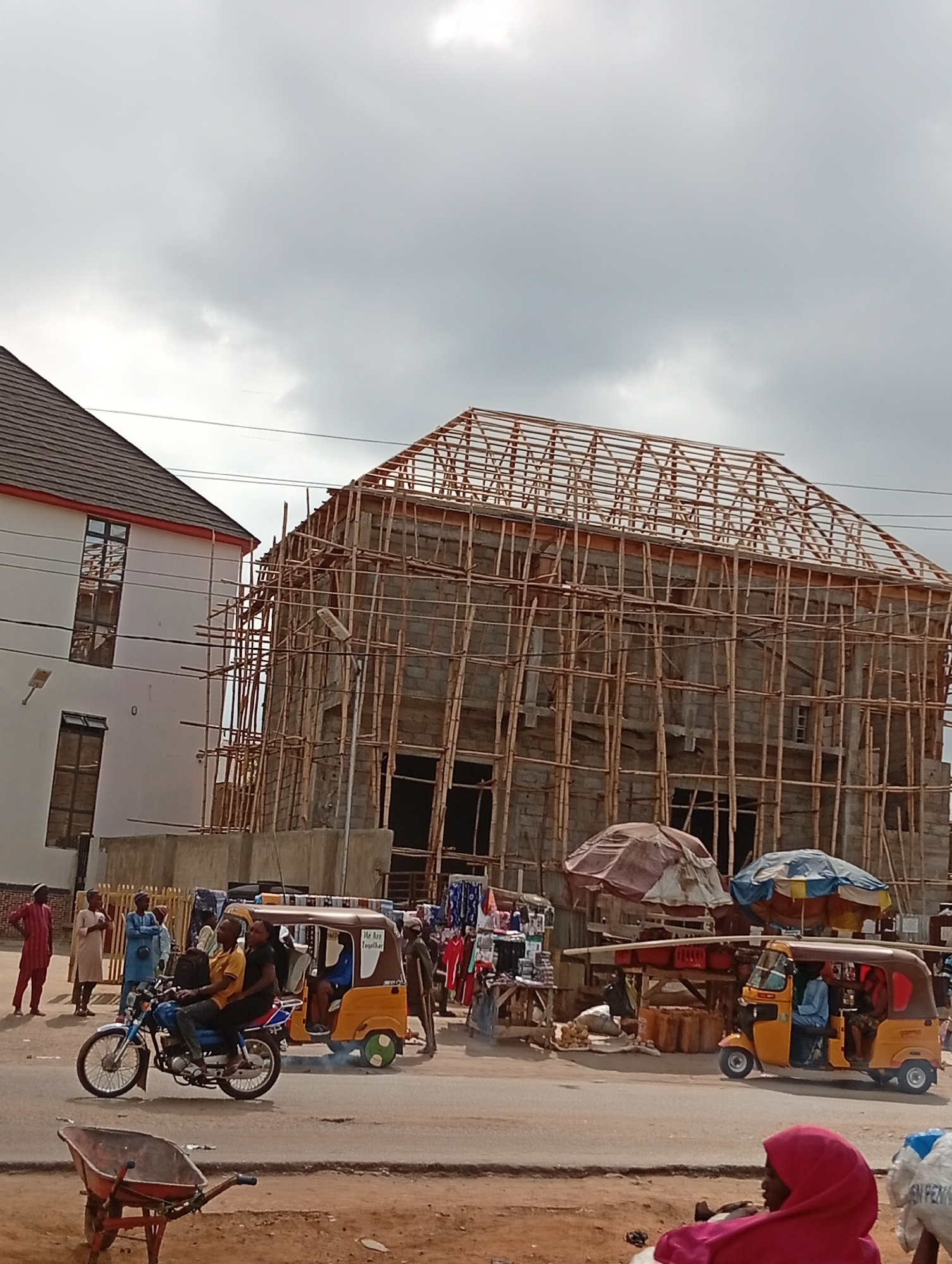 The image depicts a street scene in Masaka, Nigeria. An unfinished building with extensive bamboo scaffolding dominates the background. To the left stands a completed two-story building with windows. In the foreground, a street is visible with several vehicles: a motorcycle with two riders, and two yellow auto-rickshaws (tuk-tuks). People are standing along the roadside, and a market stall is set up with goods displayed under umbrellas. The weather appears overcast. A wheelbarrow sits in the foreground to the left.