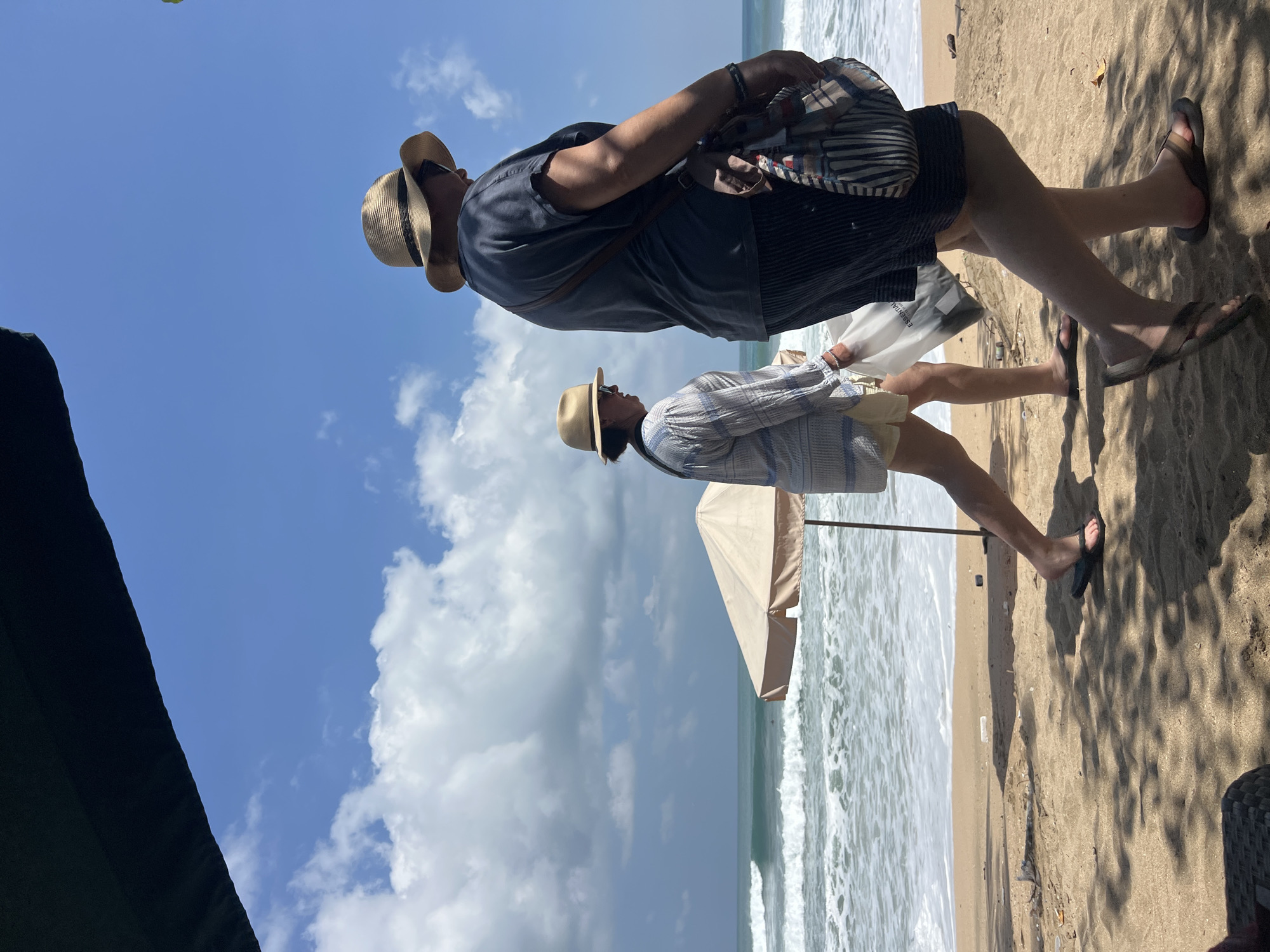 This image captures a bright, sunny day on a beach in Kuta, Indonesia. Two individuals are walking along the sandy shore near the ocean. The person in the foreground, on the right, is wearing a dark t-shirt, striped shorts, a straw hat, and flip-flops, and is carrying a colorful striped bag over their shoulder. The second person, walking to the left and slightly behind, is wearing a light-colored patterned shirt, light-colored shorts, a straw hat, and flip-flops, and is carrying a white plastic bag. Gentle waves are breaking on the shore, and a beach umbrella stands open in the background. The sky is blue with scattered white clouds, and shadows from an unseen overhead structure are cast across the sand.
