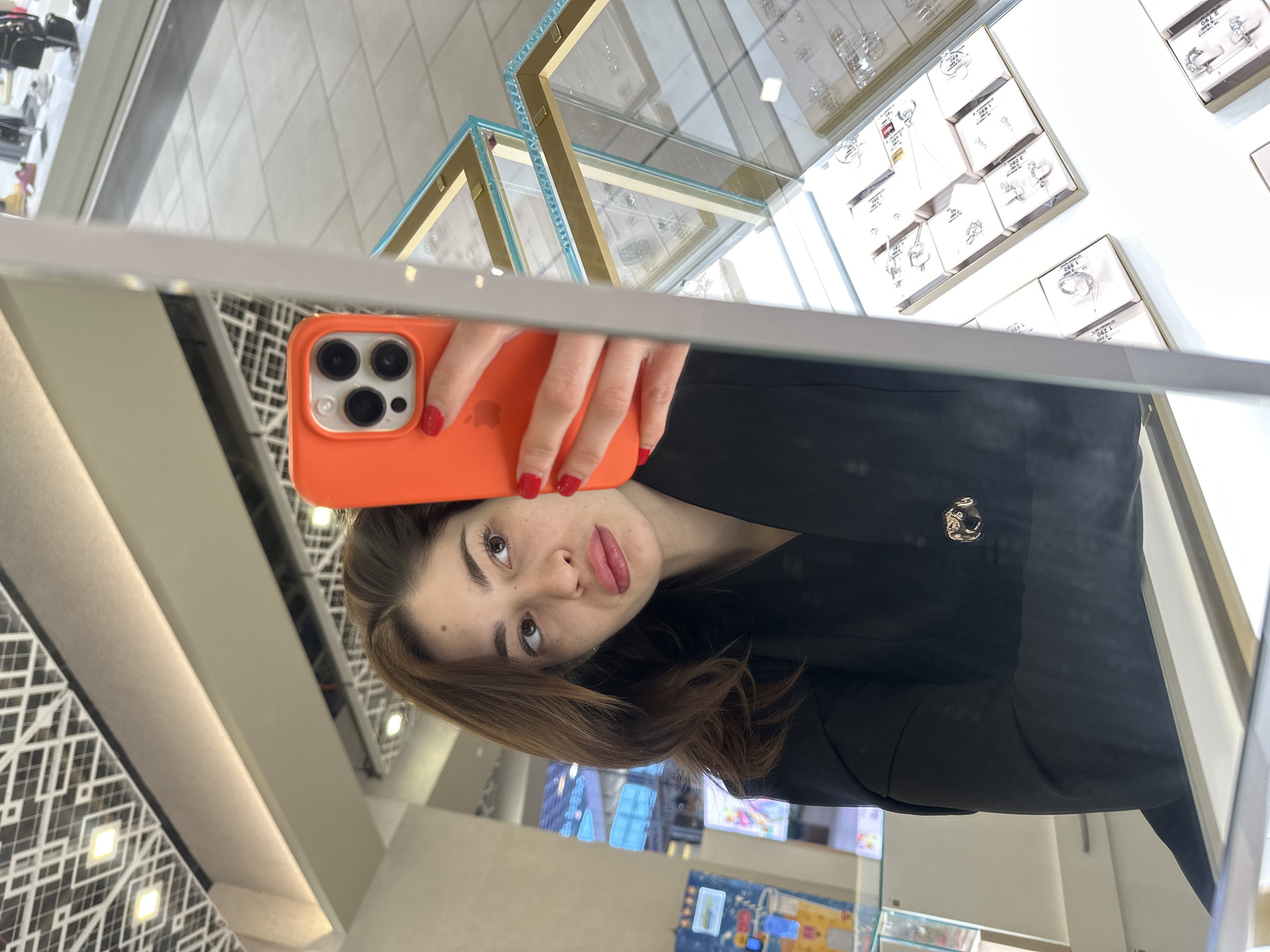 A female individual is depicted in a mirror selfie within a retail environment in Yekaterinburg, Russia, identified as a jewelry store. The subject, possessing light brown hair and a fair complexion, wears a black jacket adorned with a silver-toned animal head brooch on her left lapel. She holds an orange-cased smartphone with a triple-lens camera system in her right hand, her fingernails painted red. Her lips are slightly pursed, and her gaze is directed towards her right.
The background reflects multiple glass jewelry display cases with gold-colored trim, showcasing various rings and necklaces on light-colored stands. Price tags, such as "1 890" and "3 990", are visible on some displays. The ceiling features a white geometric pattern incorporating square and rectangular light fixtures. The floor is light-colored and tiled. Reflections in the background indicate additional retail displays and a blue-tinted architectural feature, possibly an external window.