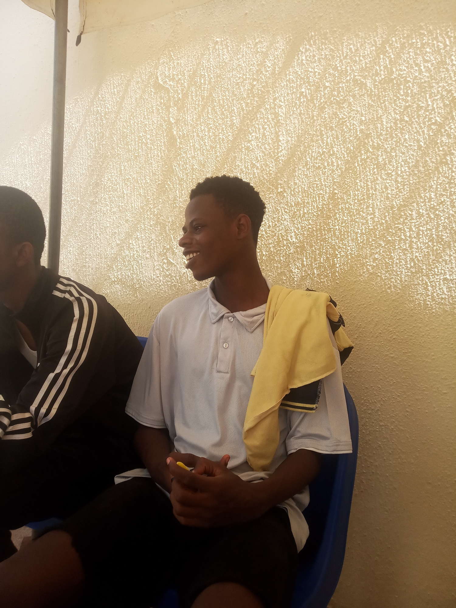 Two young men are sitting on a blue plastic chair in an indoor setting. The background features a textured, off-white wall. One man is wearing a black jacket with white stripes on the sleeves. The second man is wearing a light blue polo shirt and black shorts. He has a yellow towel draped over his shoulder and is smiling. He is holding a small yellow object in his hands. The location is Ukwu-Abwa, Nigeria.