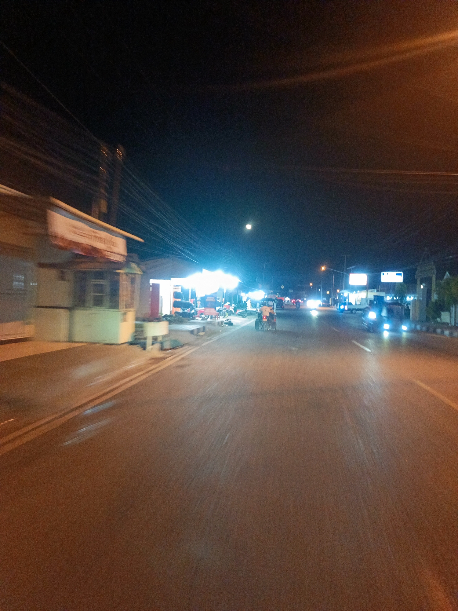 The image depicts a nighttime scene on a paved road in Ulapato A, Indonesia. The road is lined with buildings and stalls on the left side, illuminated by artificial lights. Several vehicles, including a tricycle and cars, are visible on the road, their headlights and taillights creating streaks of light due to motion blur. There are also streetlights and overhead power lines contributing to the ambient lighting. The sky is dark, with a faint illumination suggesting a moon or distant light source. The overall impression is one of a busy street scene at night.