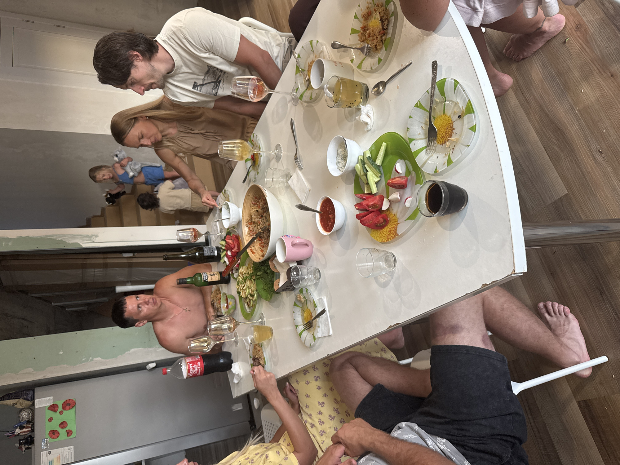 The photograph captures a gathering of people seated around a white table, likely in a dining area or a communal space. The setting appears to be an interior with a refrigerator visible on the left, adorned with decorative magnets. The overall ambiance suggests a meal or social event in progress.
At the table, six individuals are partially visible, engaged in eating and interacting. A man, shirtless and wearing a necklace, is seated on the left side of the frame, looking to the side. Next to him, a woman with blonde hair, wearing a beige dress, is leaning forward, appearing to interact with the food on the table. To her right, a man with dark hair, wearing a t-shirt, is looking towards the woman. In the background, on a staircase, two young children are visible, one in blue shorts and the other in a beige outfit.
The table is laden with food and drinks. Various dishes, including salads, rice dishes, and bowls of what appears to be sauce or dip, are spread across the surface. Several glasses, some containing drinks, are also present. A bottle of Coca-Cola and two bottles of wine are among the beverages. Plates with remnants of food and used cutlery indicate that the meal is underway or has recently concluded. The plates themselves are decorated with a floral pattern.
The flooring is made of wooden planks, and the walls exhibit unfinished construction elements, suggesting the location might be undergoing renovation or is a rustic dwelling. The lighting is ambient, with no single dominant light source. The image is taken from a slightly elevated angle, looking down at the table. The overall scene conveys a casual, familial gathering. The location is identified as Verkhniy Yurt, Russia.